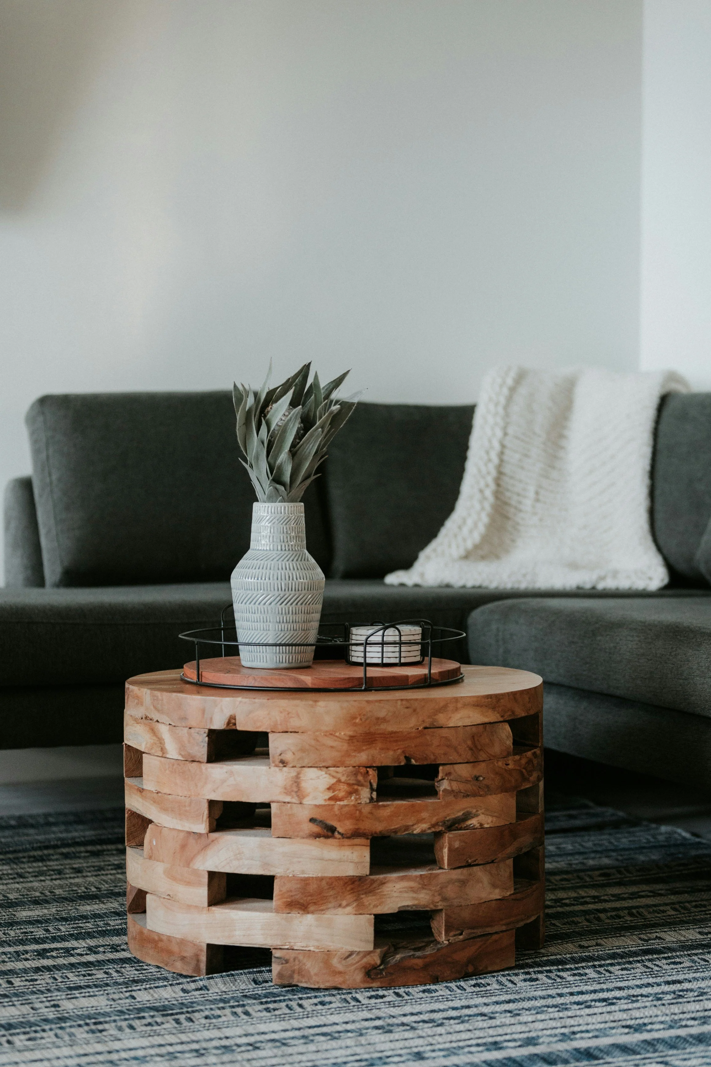 A living room with a dark gray sectional sofa, a white throw blanket, a wooden coffee table with a decorative vase and tray on top, and a patterned rug.