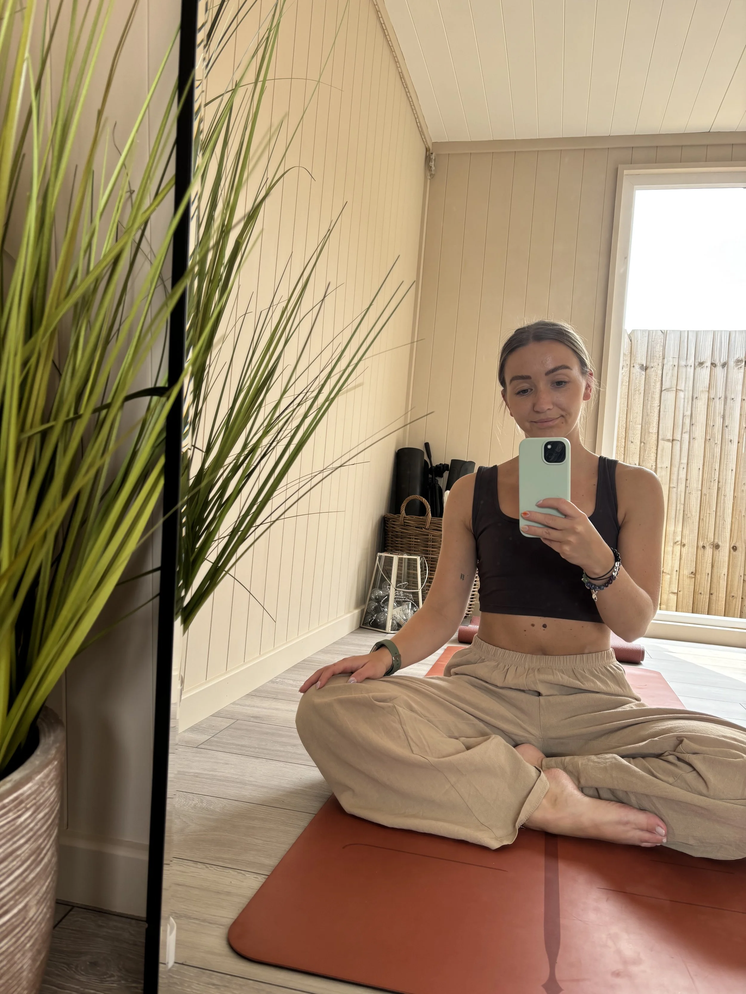A young woman sitting cross-legged on an orange yoga mat in a room with wooden floors and walls, taking a selfie in a mirror, with green plants and a wooden fence visible outside through a window.