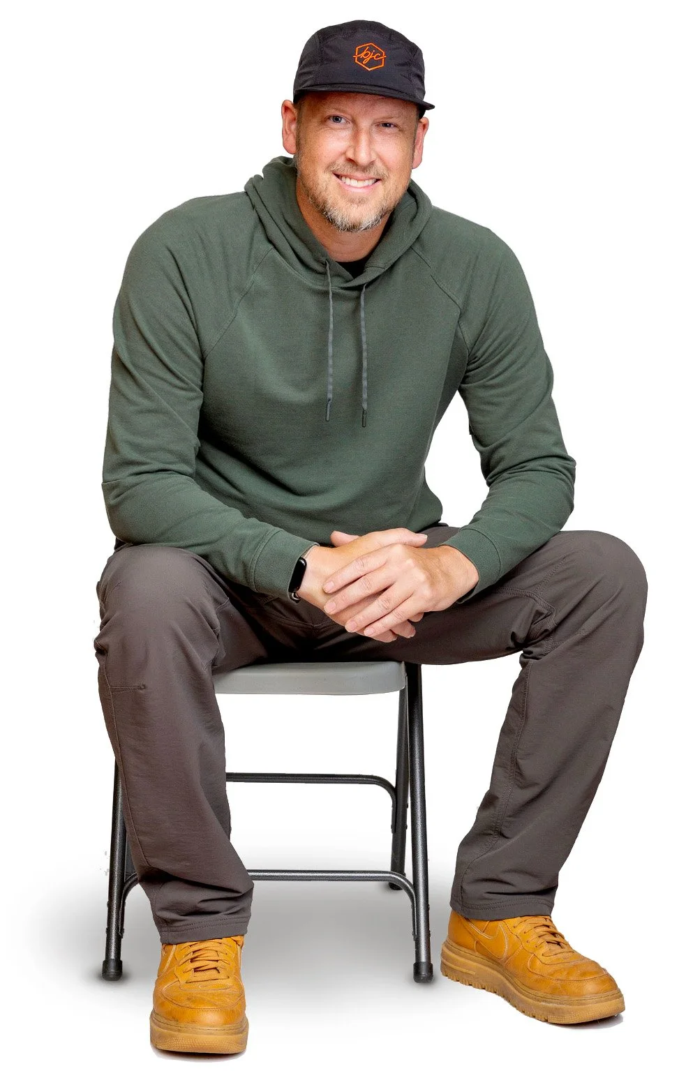 A smiling man sitting on a chair, wearing a black cap, green hoodie, brown pants, and tan boots, against a white background.
