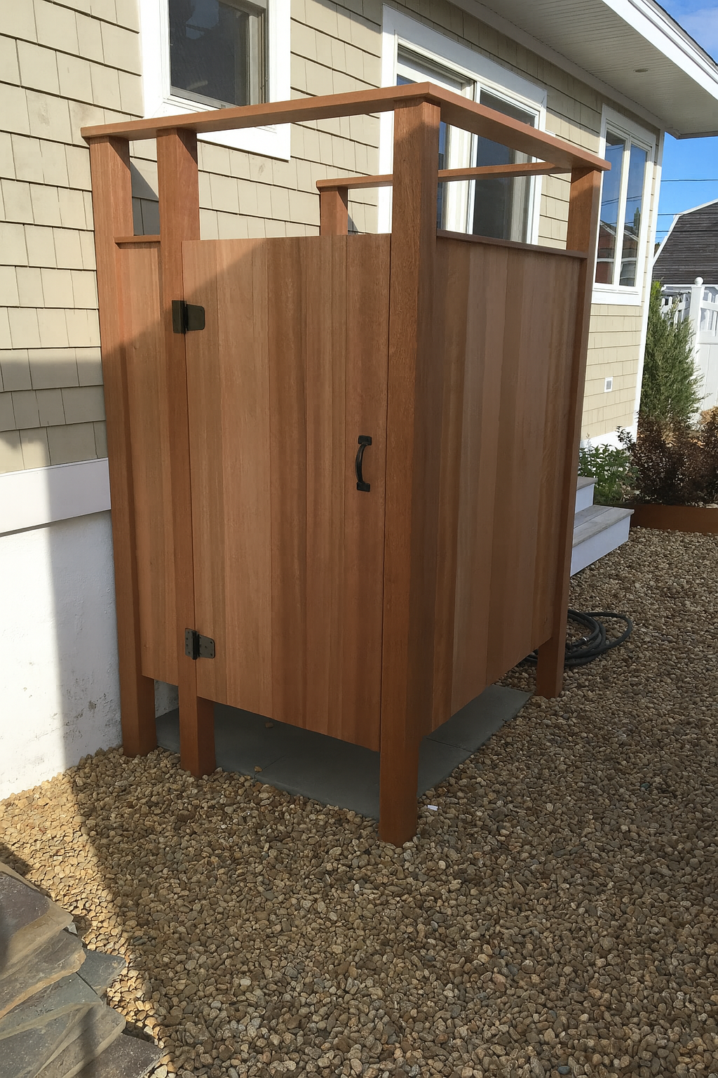 A wooden outdoor cabinet with a roof, placed on a concrete slab, outside a house with beige siding and gravel ground.