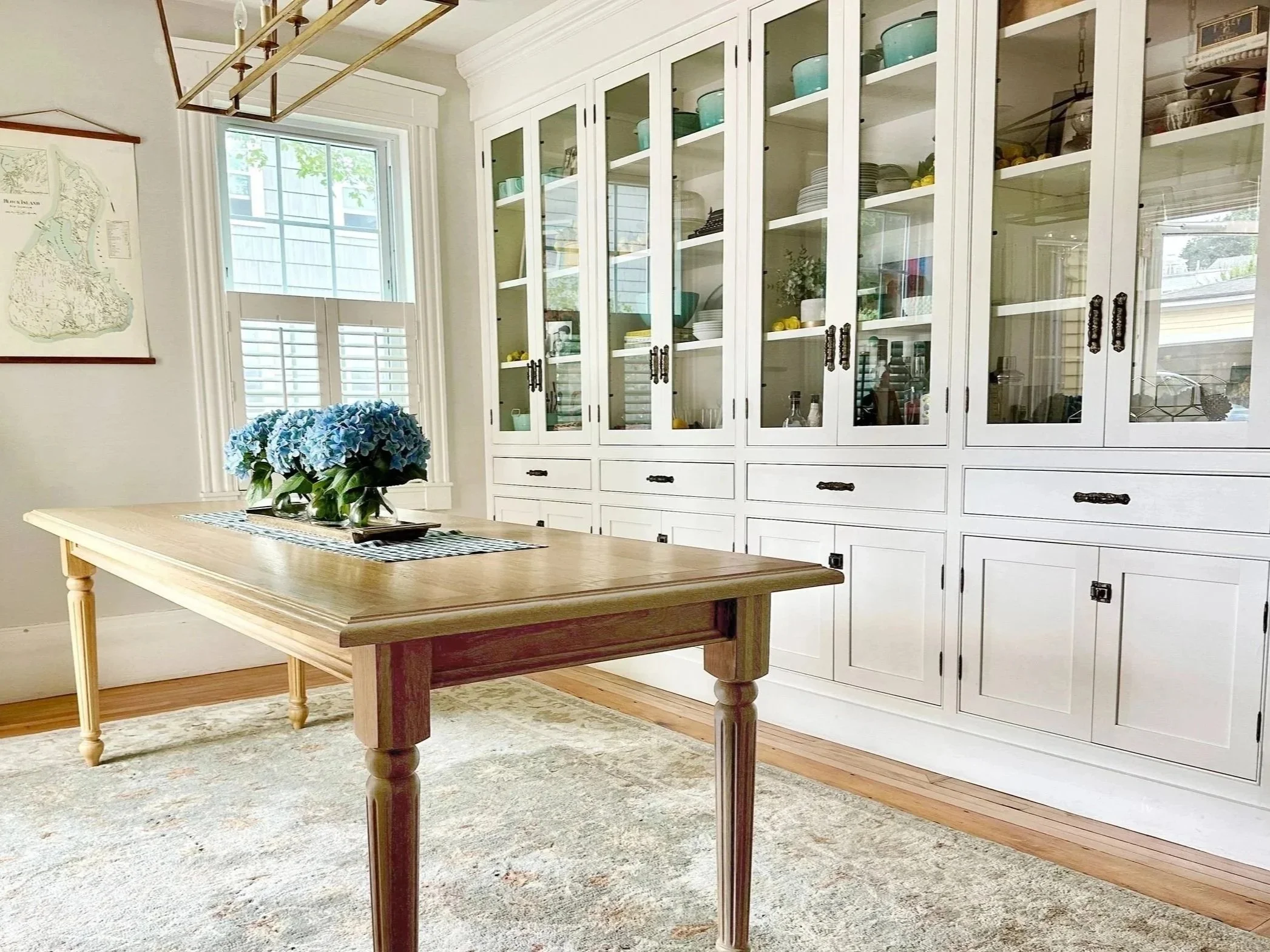 A dining room with a rectangular wooden table, six blue velvet chairs, a chandelier above, white built-in china cabinet, a window with shutters, and a bouquet of yellow flowers on the table.