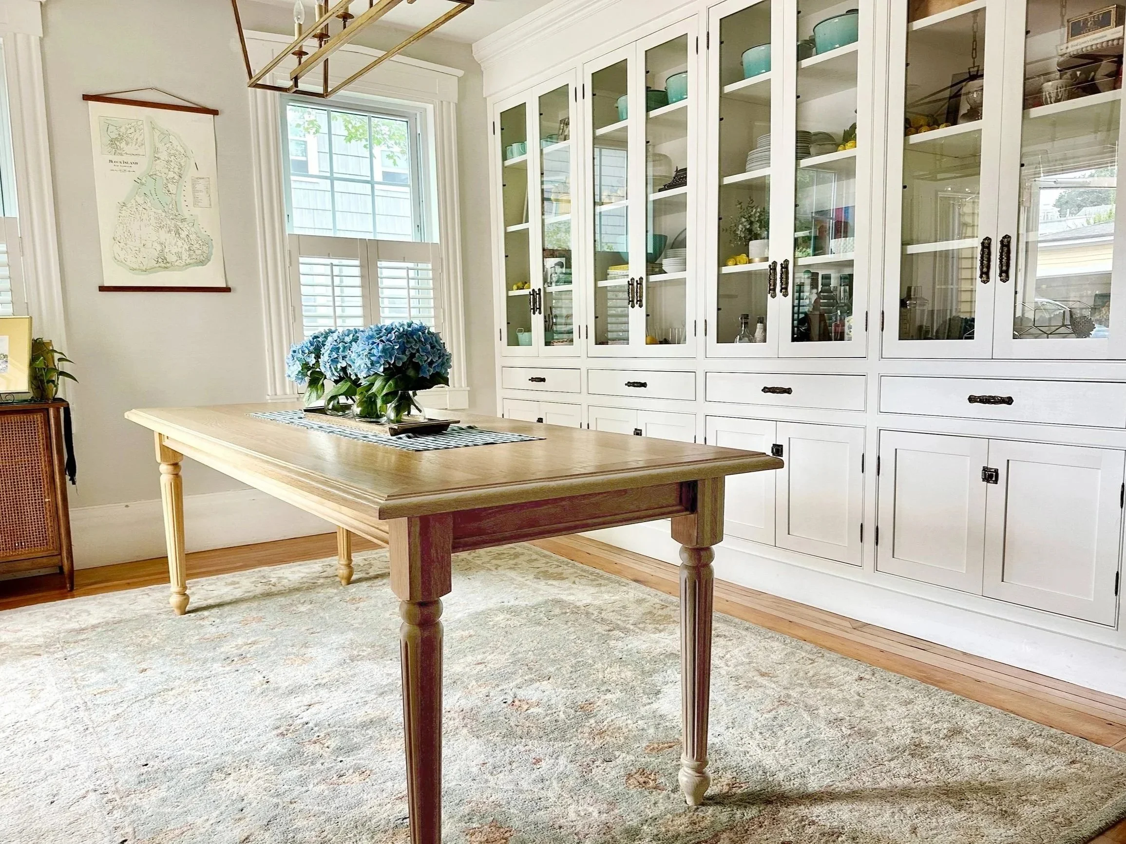 Custom dining room built-in white cabinet, with custom white oak table, blue floral centerpiece, and large window with shutters.