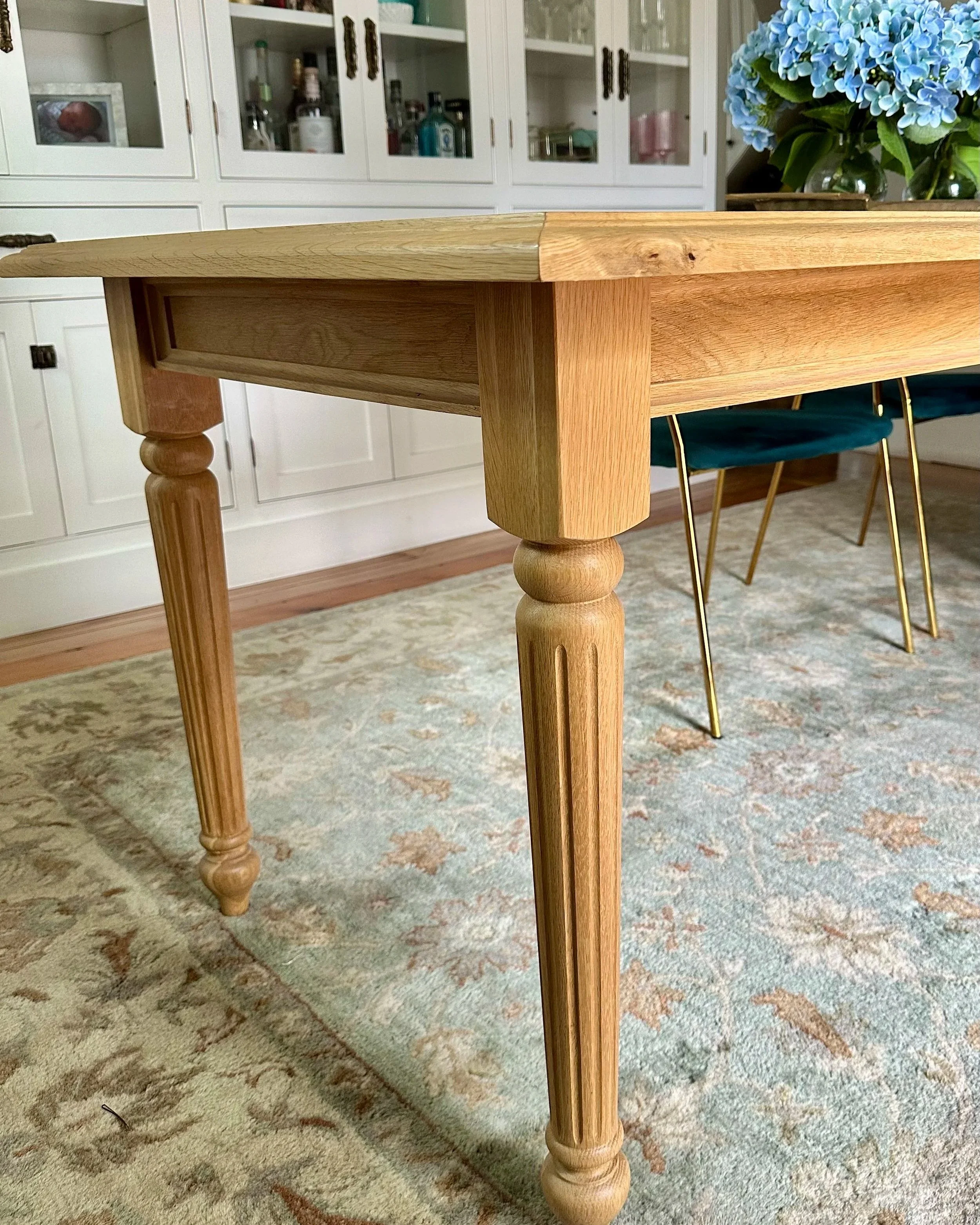 A dining room table surrounded by a mix of vintage and modern chairs, with a vase of flowers on top, near windows with white shutters.