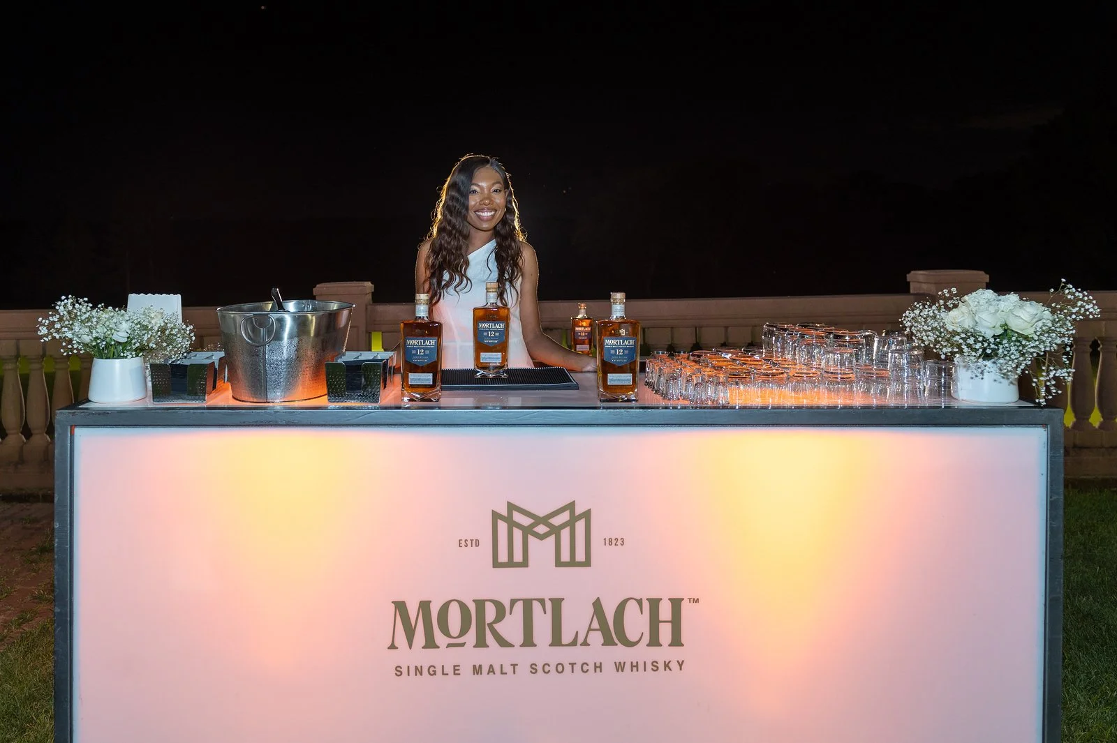 A woman stands behind an illuminated outdoor bar at night. The bar front reads 'Mortlach Single Malt Scotch Whiskey'. 