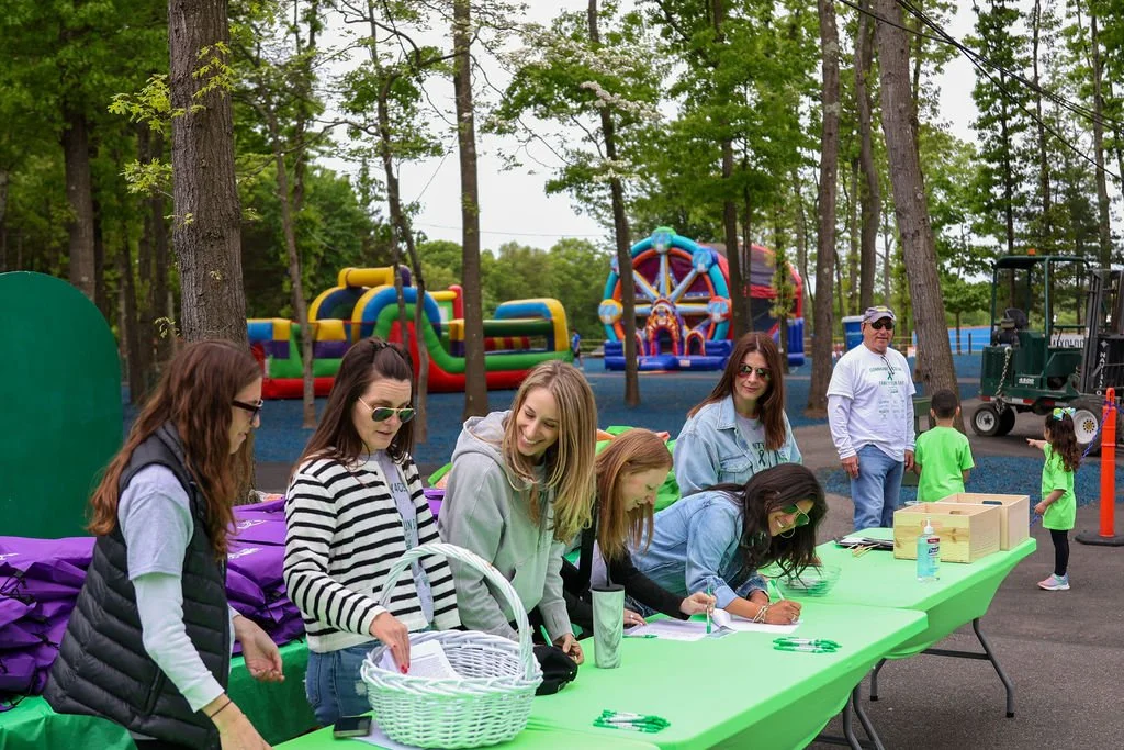 Group of women signing in at a registration table outdoors at a park, with inflatable bounce houses and children in the background.