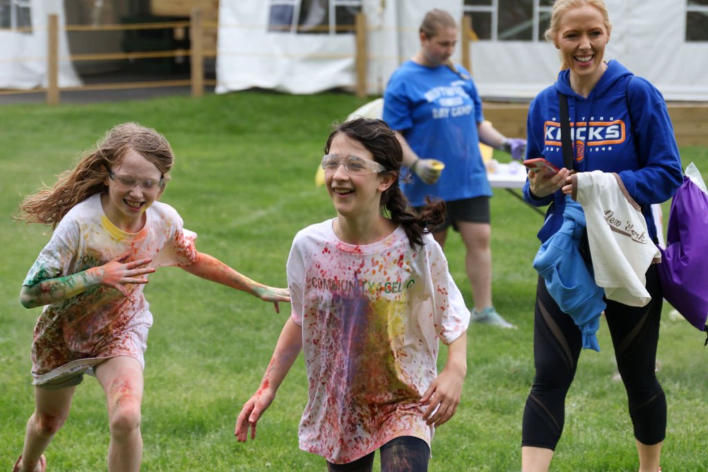 Two young girls and two adult women run outdoors, participating in a color run event. The girls are covered in colored powder, laughing and running, while the woman, smiling, stands nearby with a phone and bags.