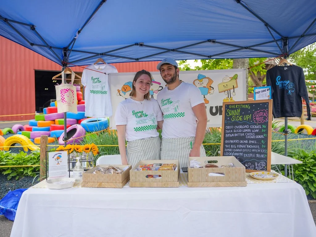 Two people standing behind a table with baked goods at a food stand, under a blue canopy, with colorful tire decorations in the background. Signage indicates gluten-free treats, including cookies, brownies, and banana bread.