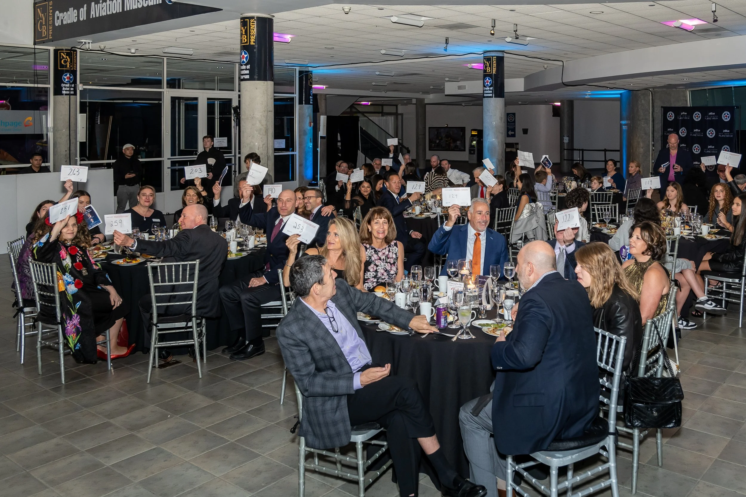 Guests are seated at round tables, raising their bidding numbers, in an event hall with dim lighting and some purple and blue ambiance.