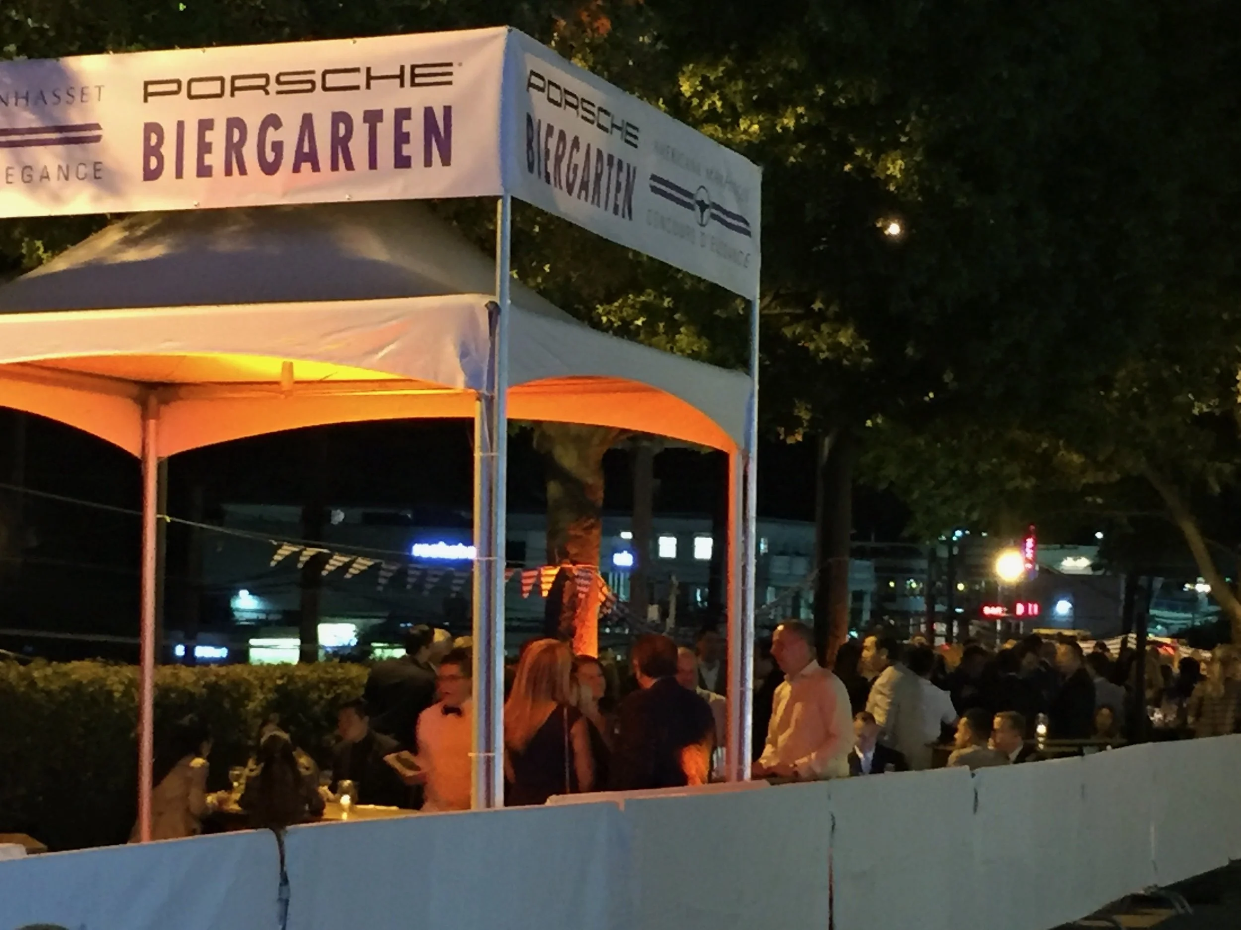 Guests socializing outside at a night event under a white tent with a sign that reads 'Porsche Biergarten'.