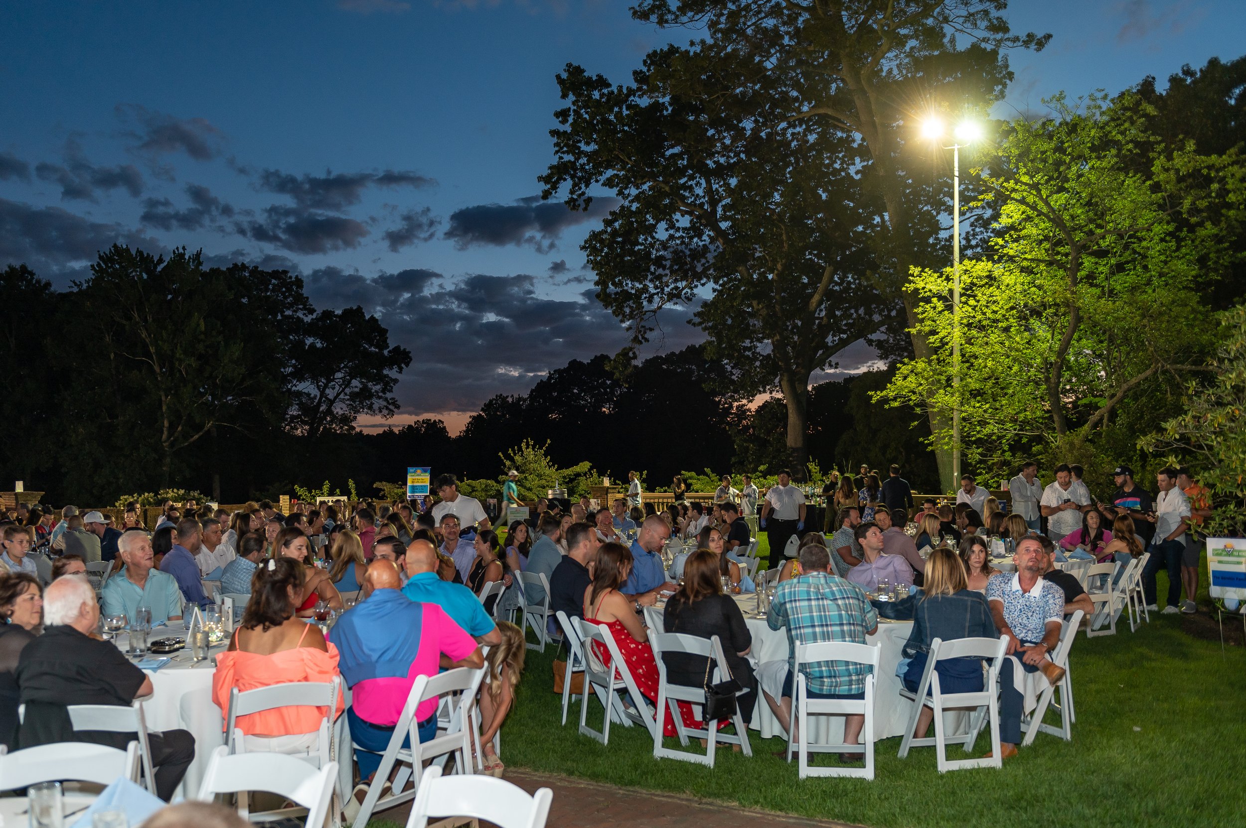 People dining outdoors at a large event during the evening, with tables and chairs set up on a grassy area, surrounded by trees and lit by overhead lights, under a darkening sky with clouds.