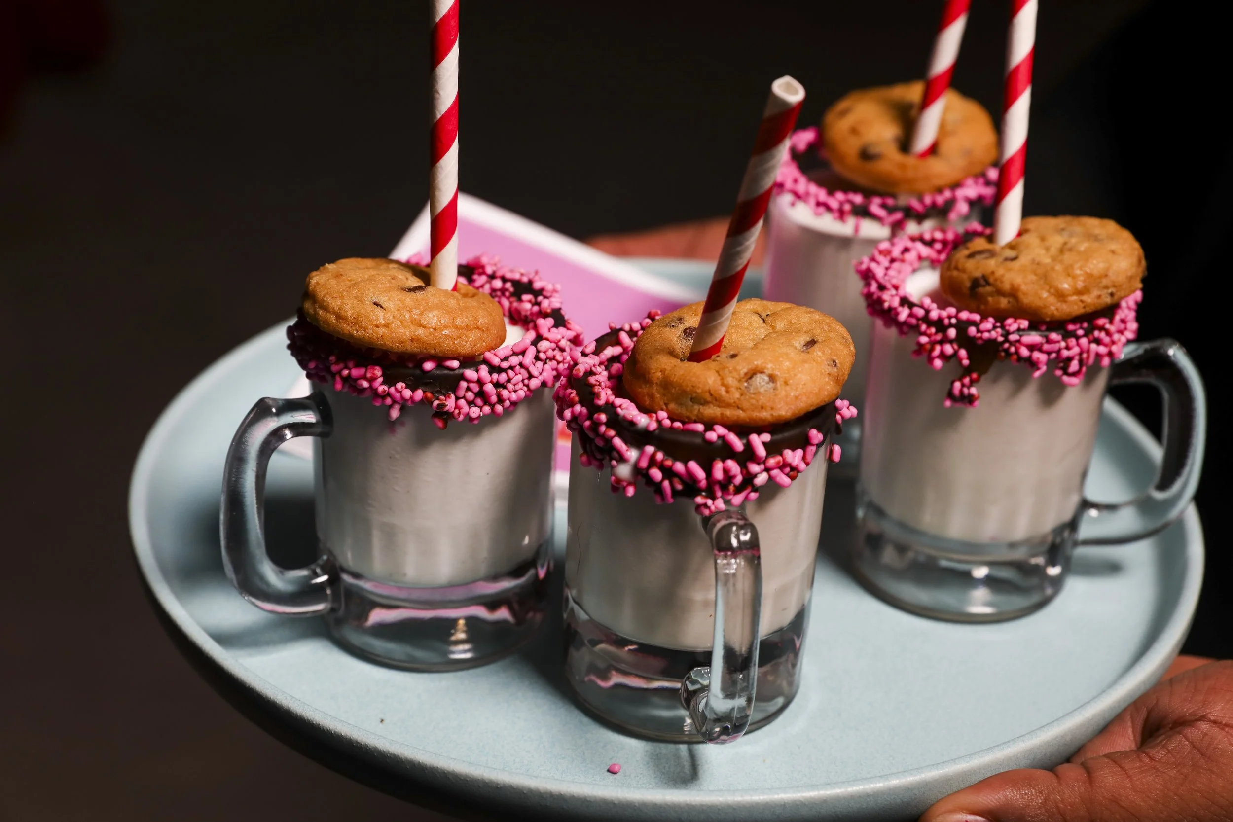 Three glass mugs filled with a creamy beverage, decorated with pink sprinkles around the rims, topped with chocolate chip cookies and pink-and-white striped straws, on a light blue tray.