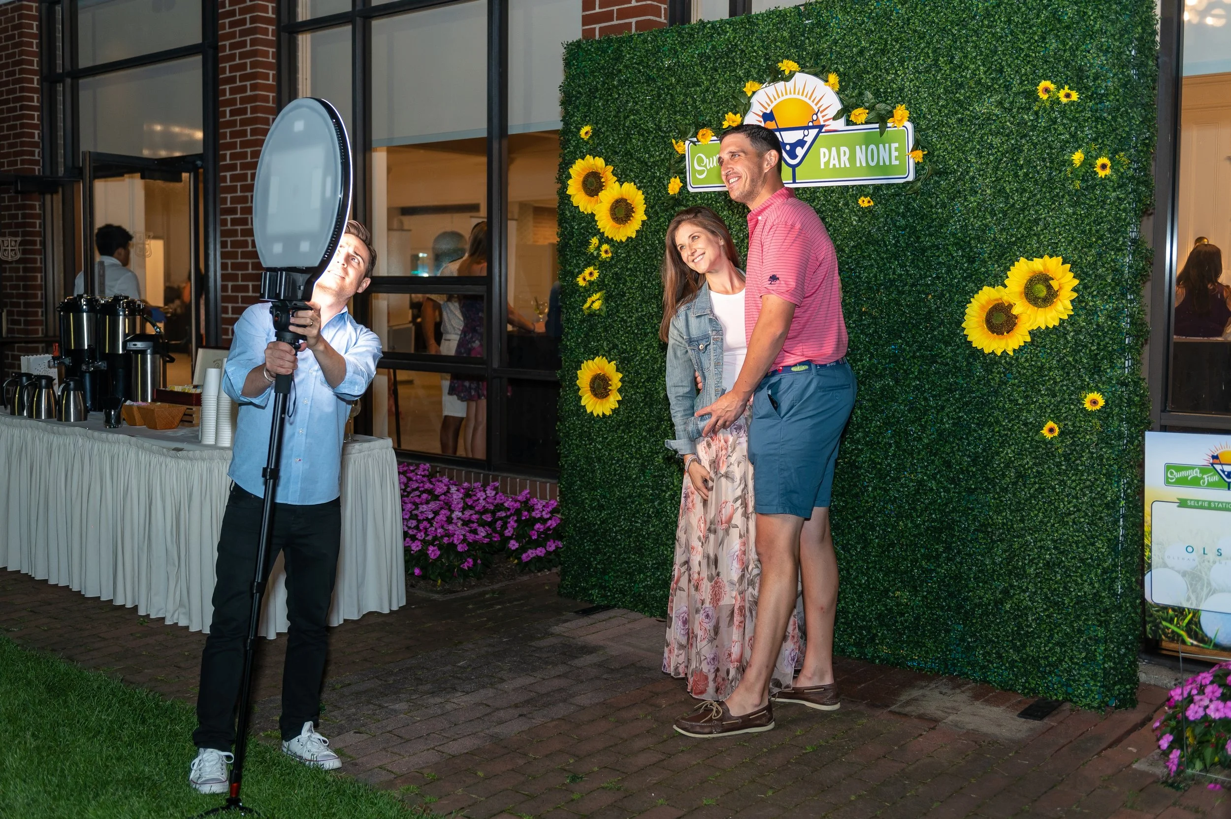 A couple getting their photo taken standing in front of a green sponsor media wall decorated with sunflowers at an outdoor event with a sign that reads 'Summer Fun Par None'.