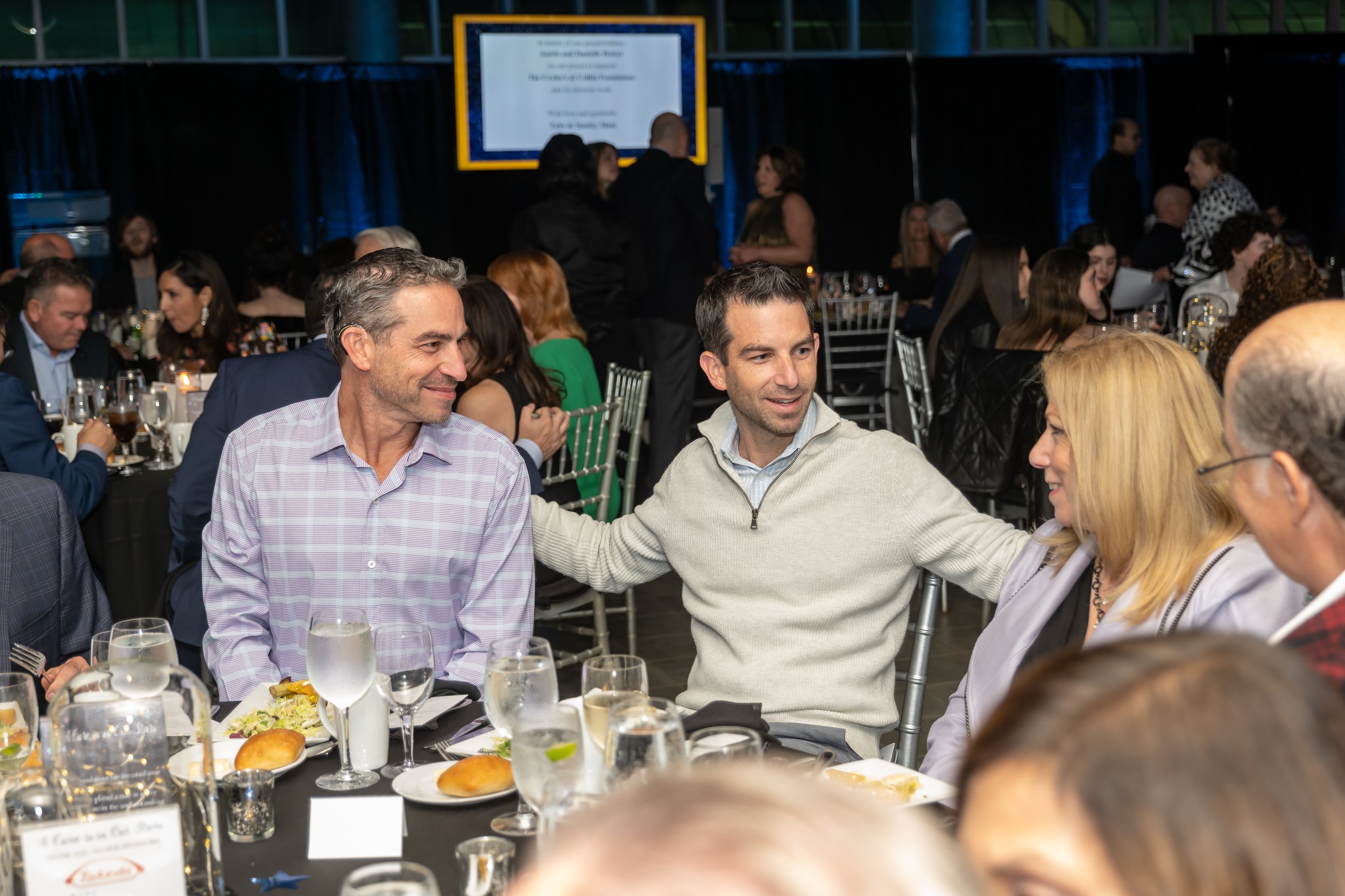 Group of people sitting at a formal dinner, engaging in conversation, with tables set with plates, glasses, and food, in a decorated event hall.