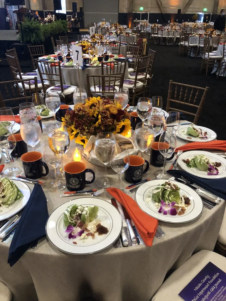 Round banquet table set for a formal event with plates of salad, wine glasses, black mugs with orange interiors, orange and navy napkins, a floral centerpiece with yellow and burgundy flowers, and a small lit candle, surrounded by empty chairs with w