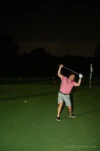 A guest on the golf course green at night raising a golf club over his head with a fun expression on his face.