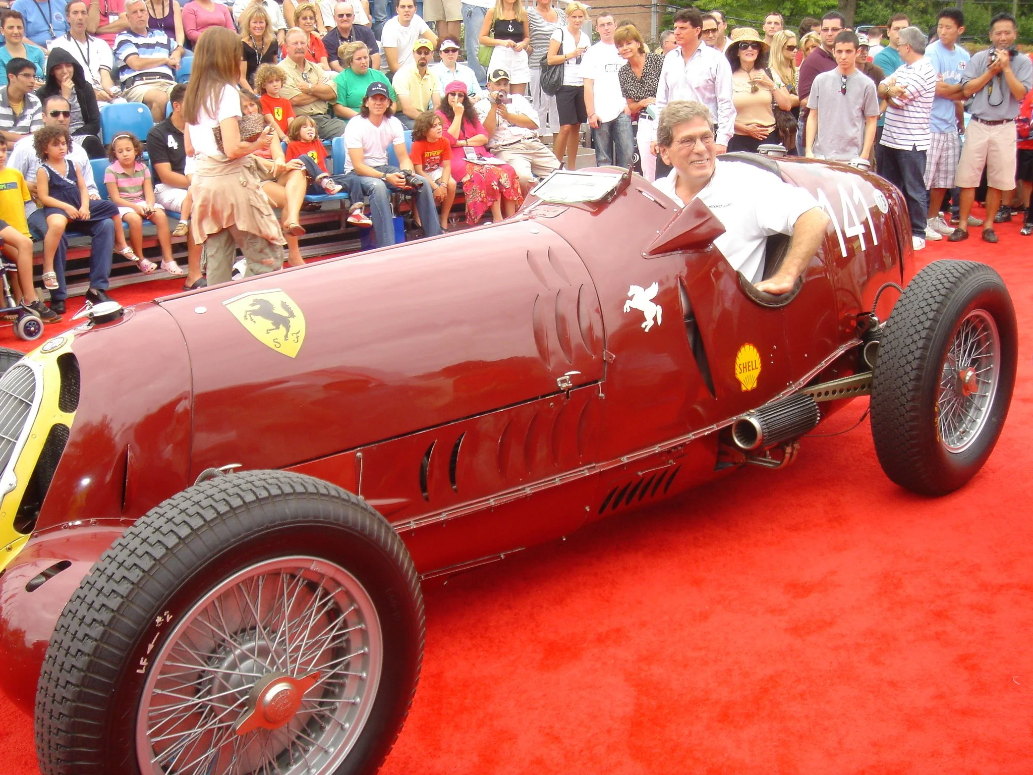 A vintage Ferrari red racing car with a man sitting in the driver's seat, surrounded by a crowd of spectators at an outdoor event.