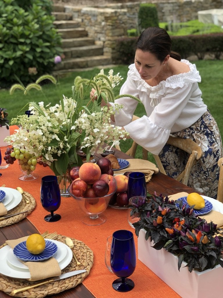 A woman in a white blouse and floral skirt arranging flowers on a table outdoor, which is decorated with fruit, flowers, and tableware for a gathering or meal.