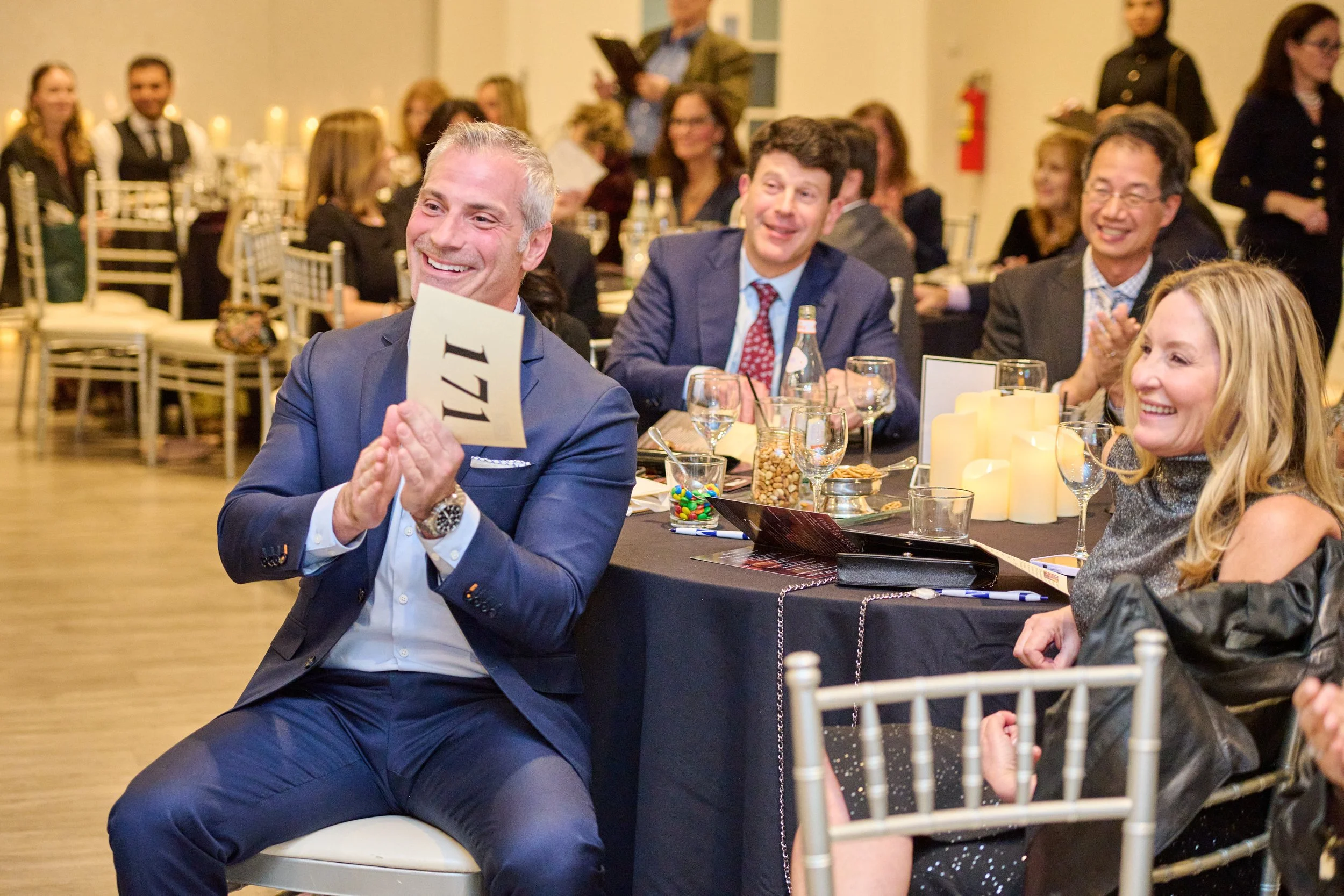 Guests at a formal event, clapping and smiling, with a man holding his bidding number 171 in the foreground.
