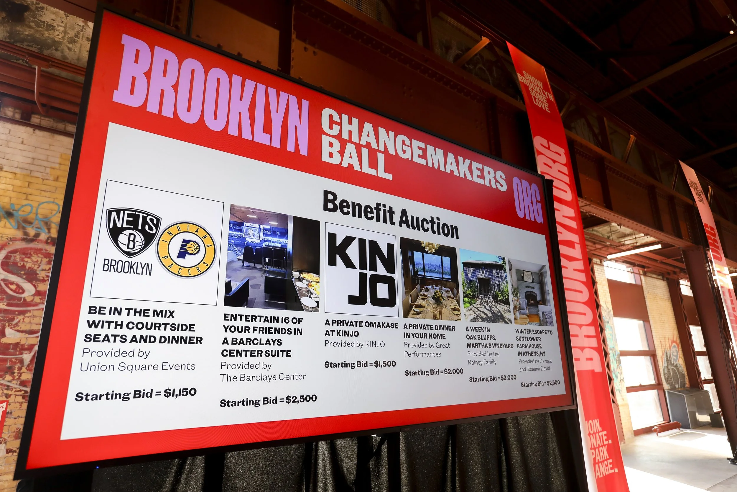 Large digital screen displaying graphics and text that read: Brooklyn Changemakers Ball benefit auction with logos of Brooklyn Nets and Indiana Pacers, and details of auction items and bids. The screen is in an industrial-style space with brick walls