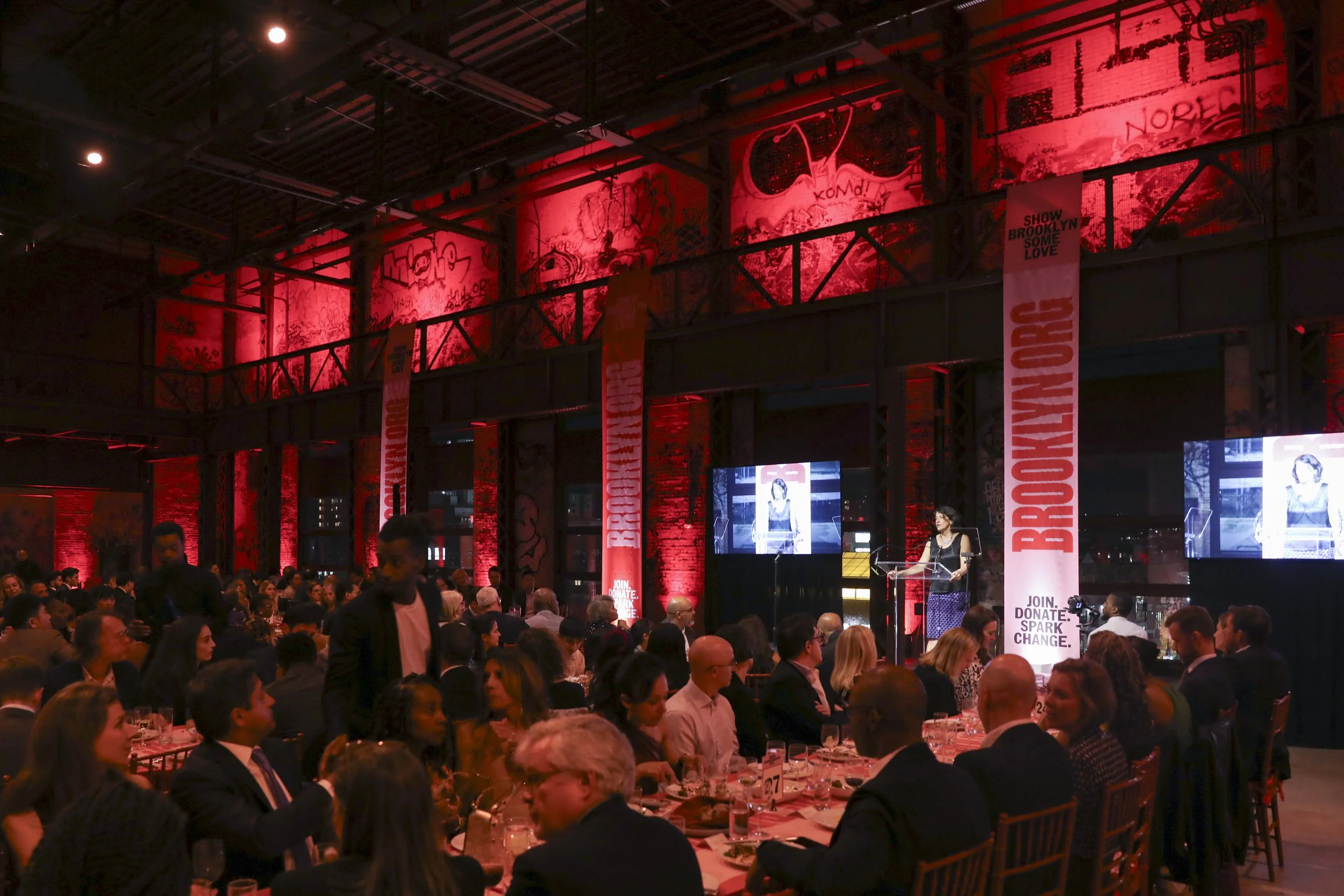 An indoor event with a large seated audience, a woman speaking at a podium on stage, multiple screens displaying her image, and red banners with 'Brooklyn Org' written on them, in a venue with graffiti-style art backdrops.