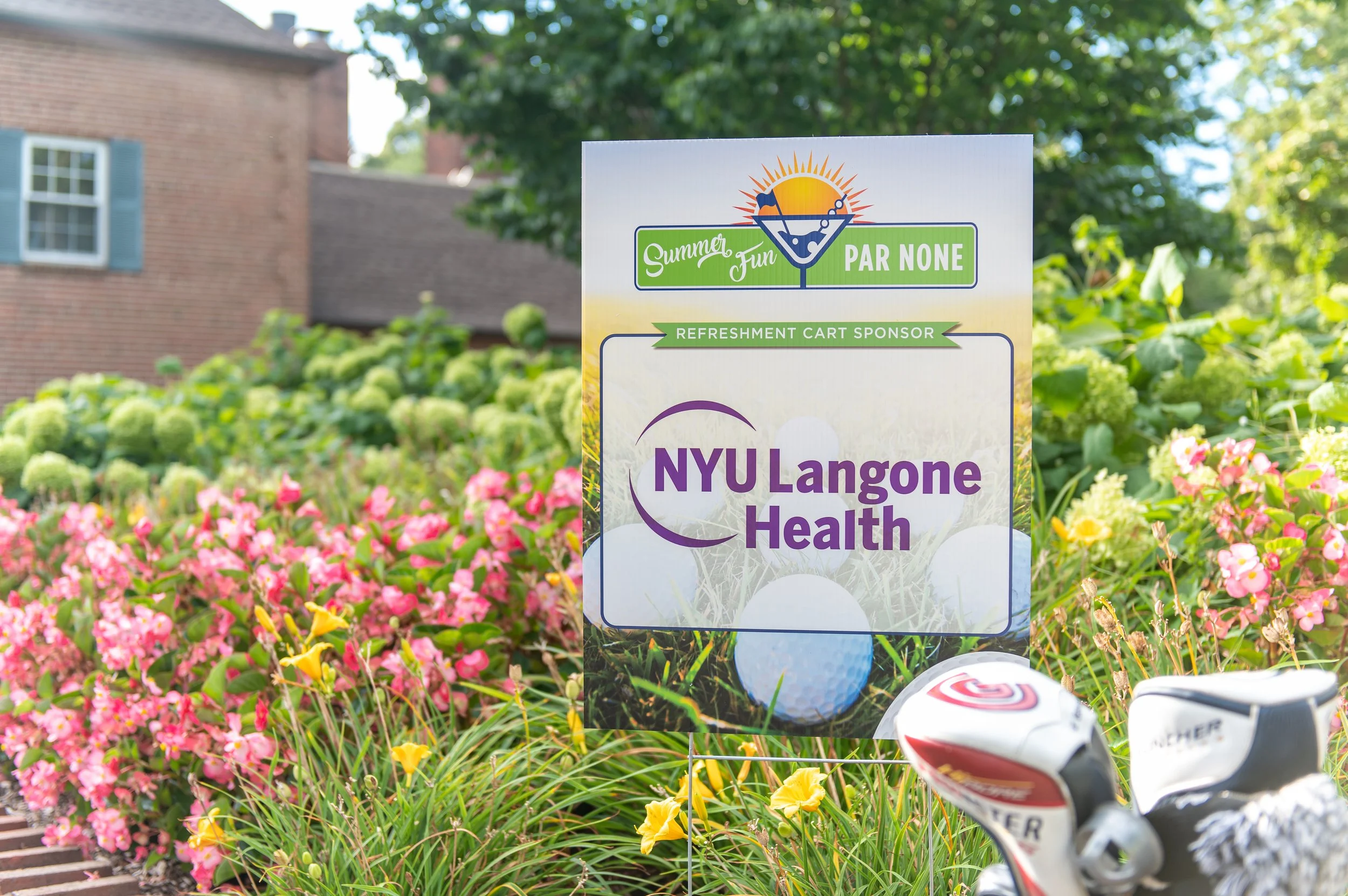 Sign reading "NYU Langone Health" in a garden with pink and yellow flowers, with golf clubs in foreground.