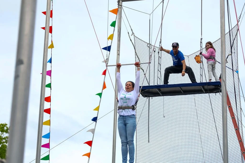 A woman swings on a trapeze while a man and a young girl stand behind her on a platform. Colorful flags decorate the poles around the course.