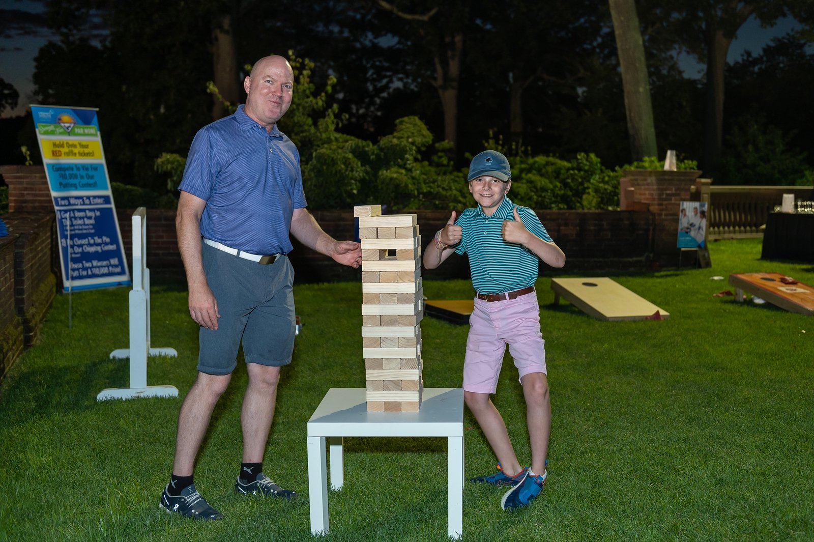 A man and a young boy give the thumbs up with a stacked giant Jenga puzzle on a table in the foreground at night.