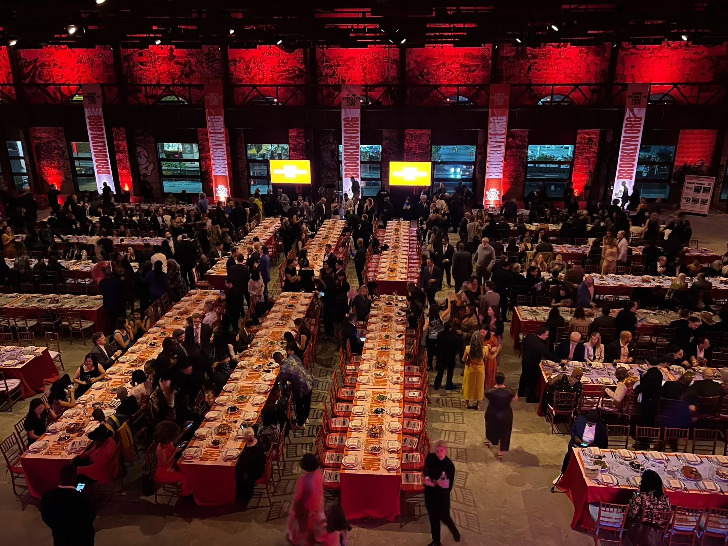 A large indoor banquet hall filled with numerous long tables set with place settings for a dinner event, with many guests seated and mingling. The background features red lighting with graffiti-style artwork on the walls and multiple large screens showing yellow and black logos. The atmosphere appears lively and social.