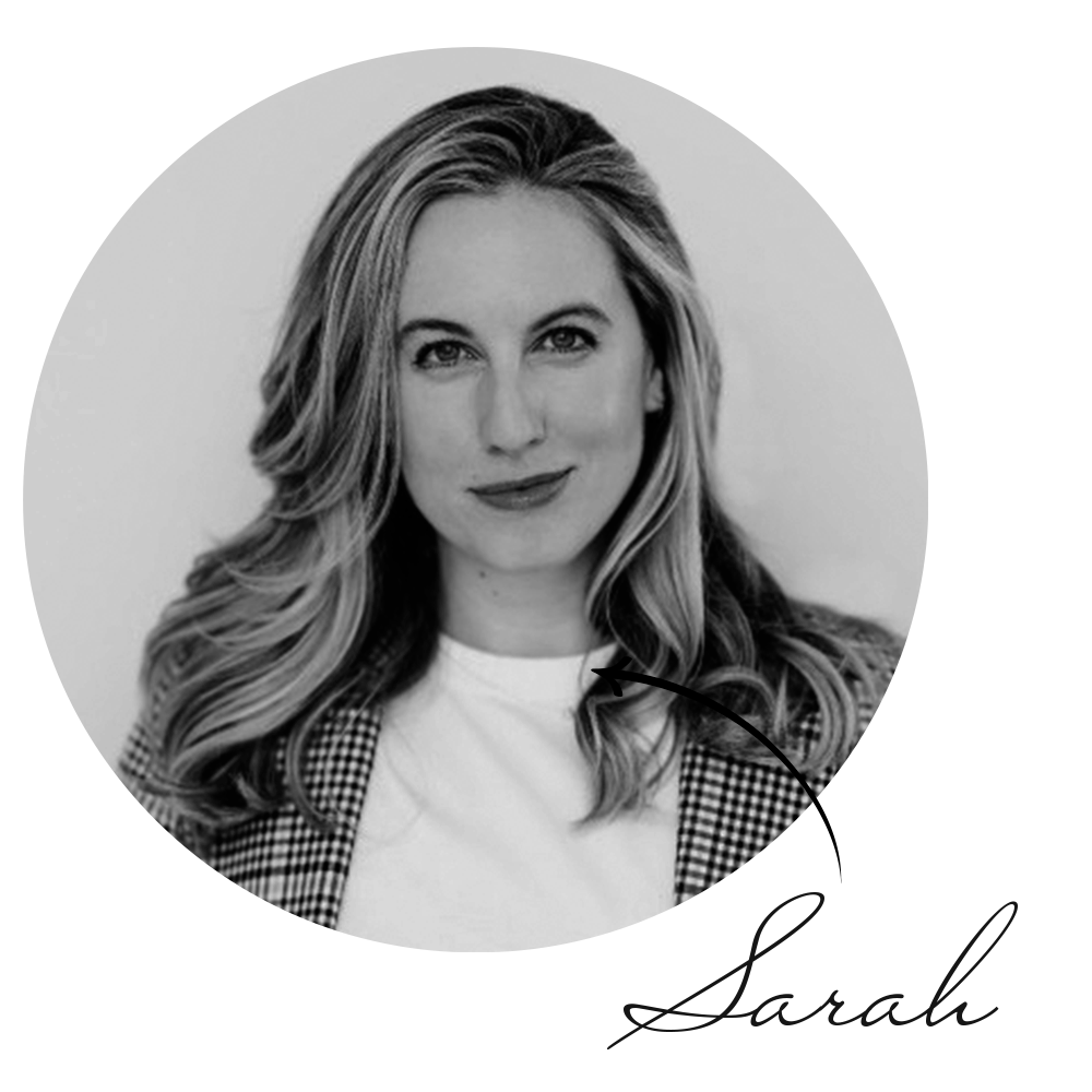 A black and white photo of a woman with long wavy hair, wearing a checkered blazer and a white top, smiling at the camera.