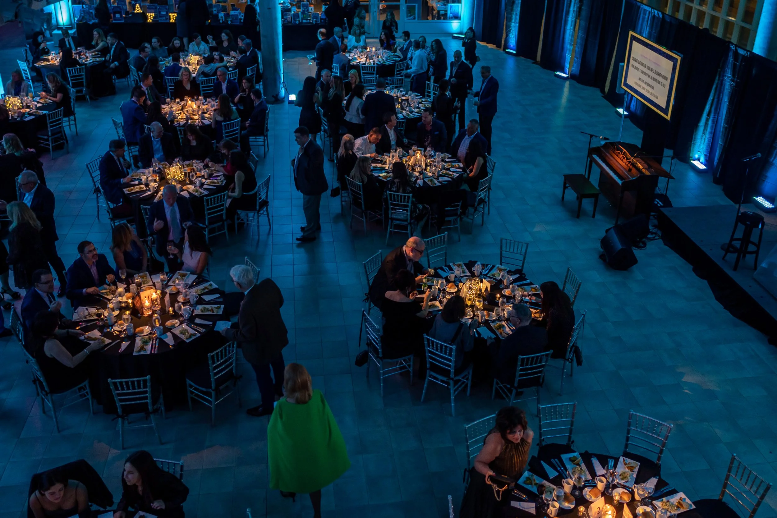 People attending a formal dinner event in a large, dimly-lit hall with round tables, candlelit centerpieces, and a stage with a screen and piano.