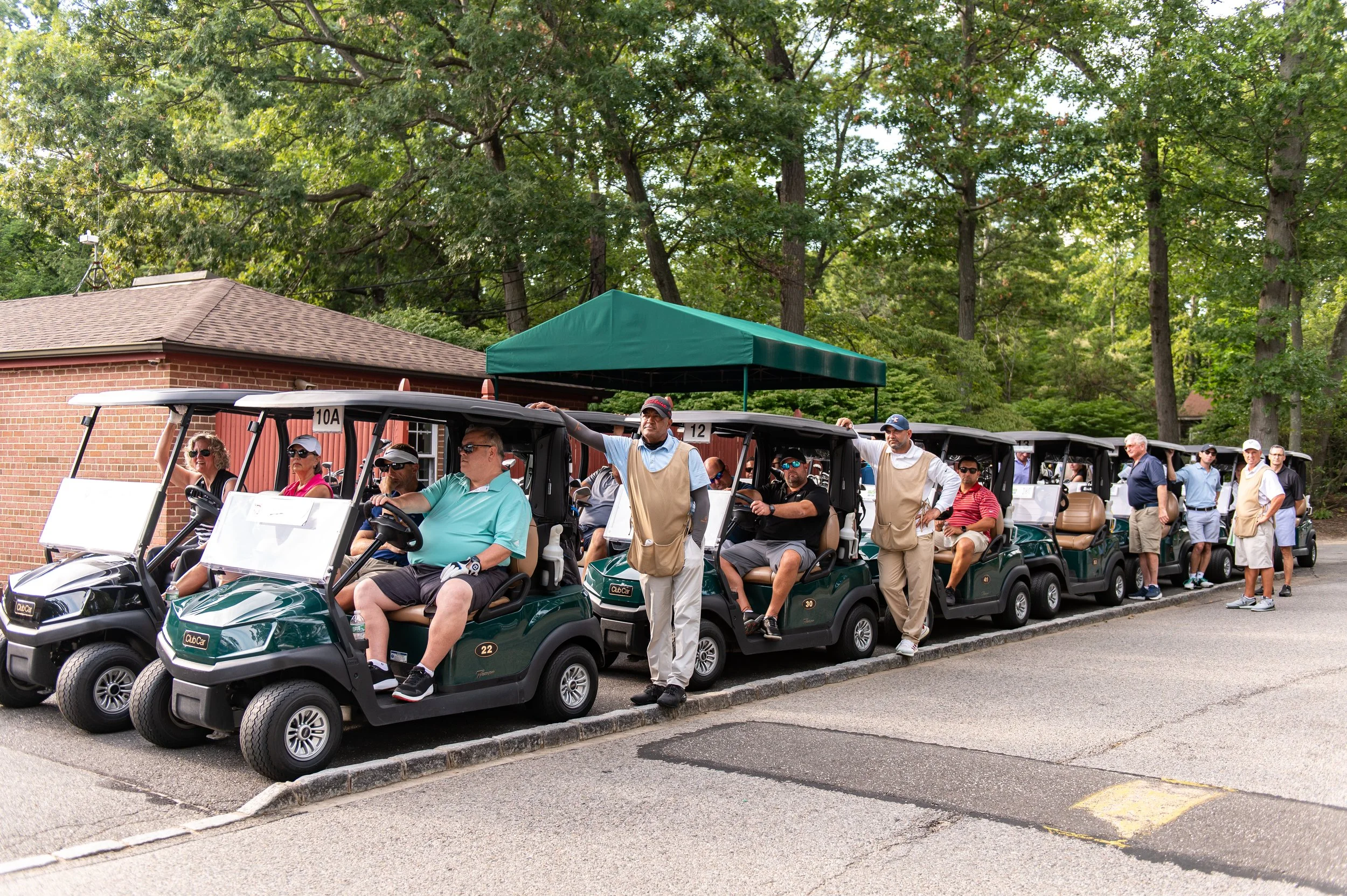 Line of people in golf carts parked outside, with a green canopy and trees in the background.