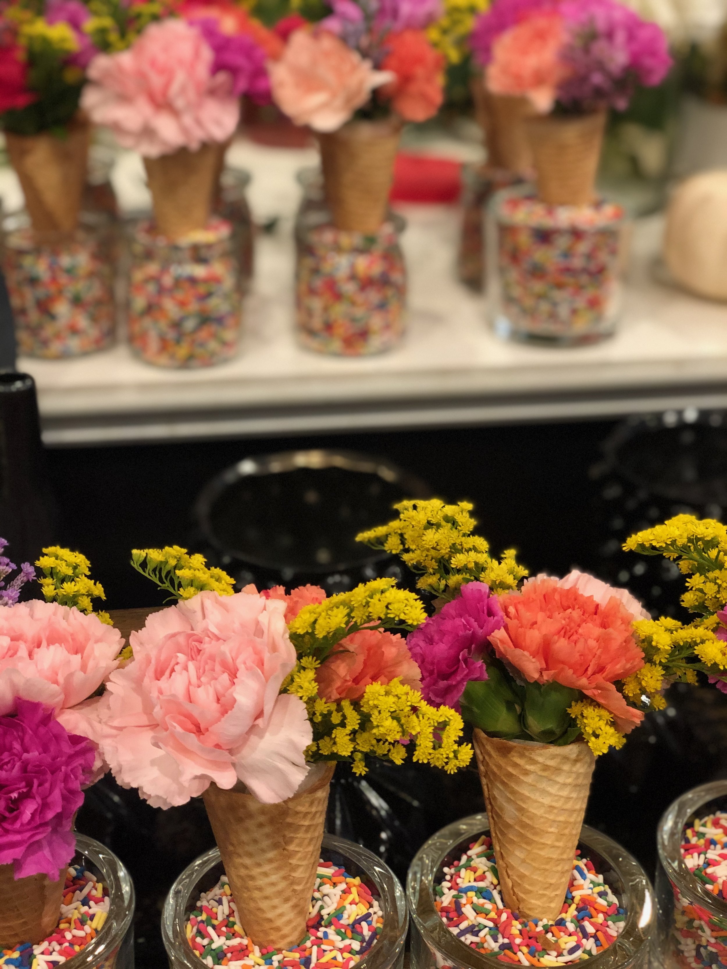 Colorful flower arrangements in waffle cone holders with sprinkles, displayed in small glass jars.