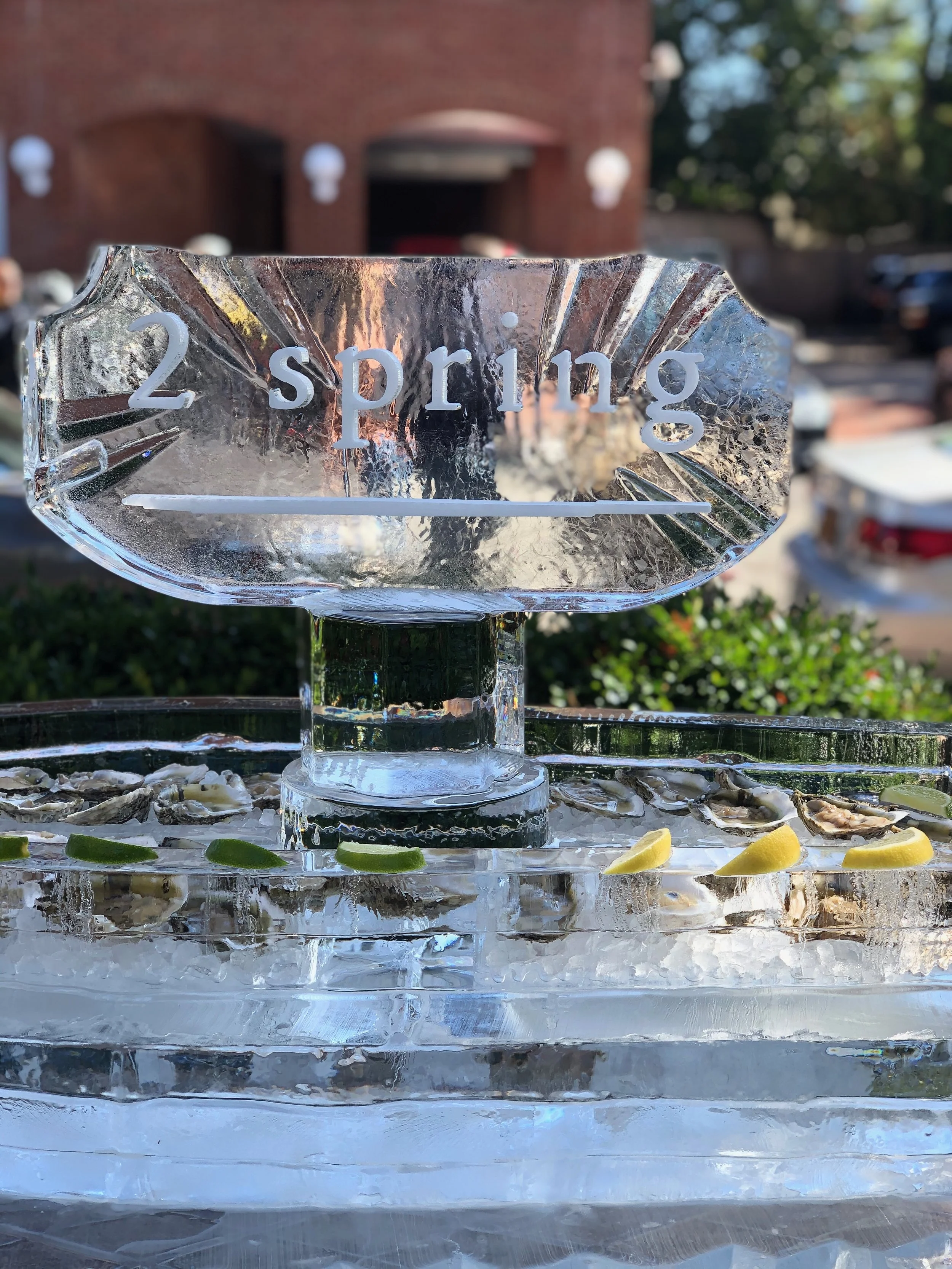 Close-up of an ice sculpture with the inscription '2 spring' on it, surrounded by garnishes like oysters and lemon slices, set outdoors with a blurred background of a brick building, trees, and parked cars.