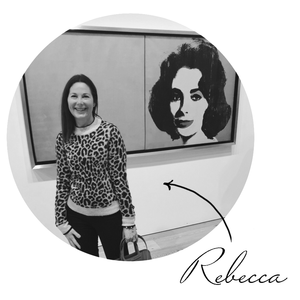 A woman smiling at an art gallery stands in front of a large black-and-white portrait painting of a woman with dark hair. The woman is wearing a leopard print sweater and holding a handbag.