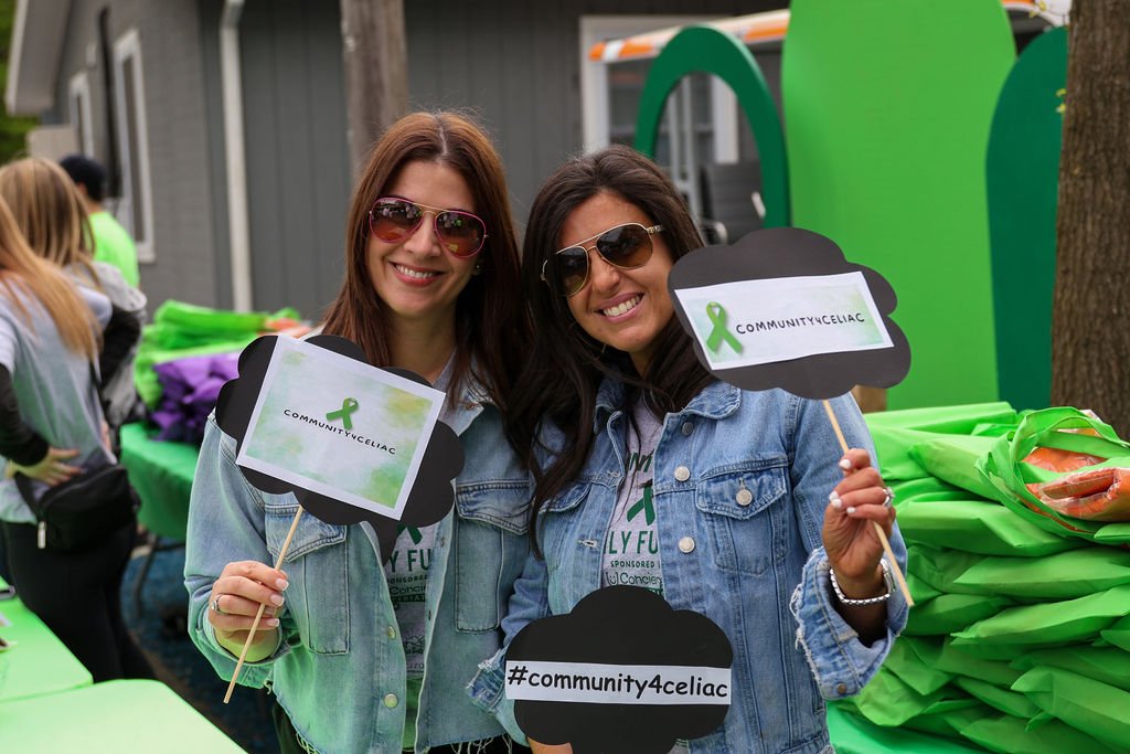 Two women smiling, wearing sunglasses and denim jackets, holding signs with green awareness ribbons and hashtags, at a community event with green bags and a green mural in the background.