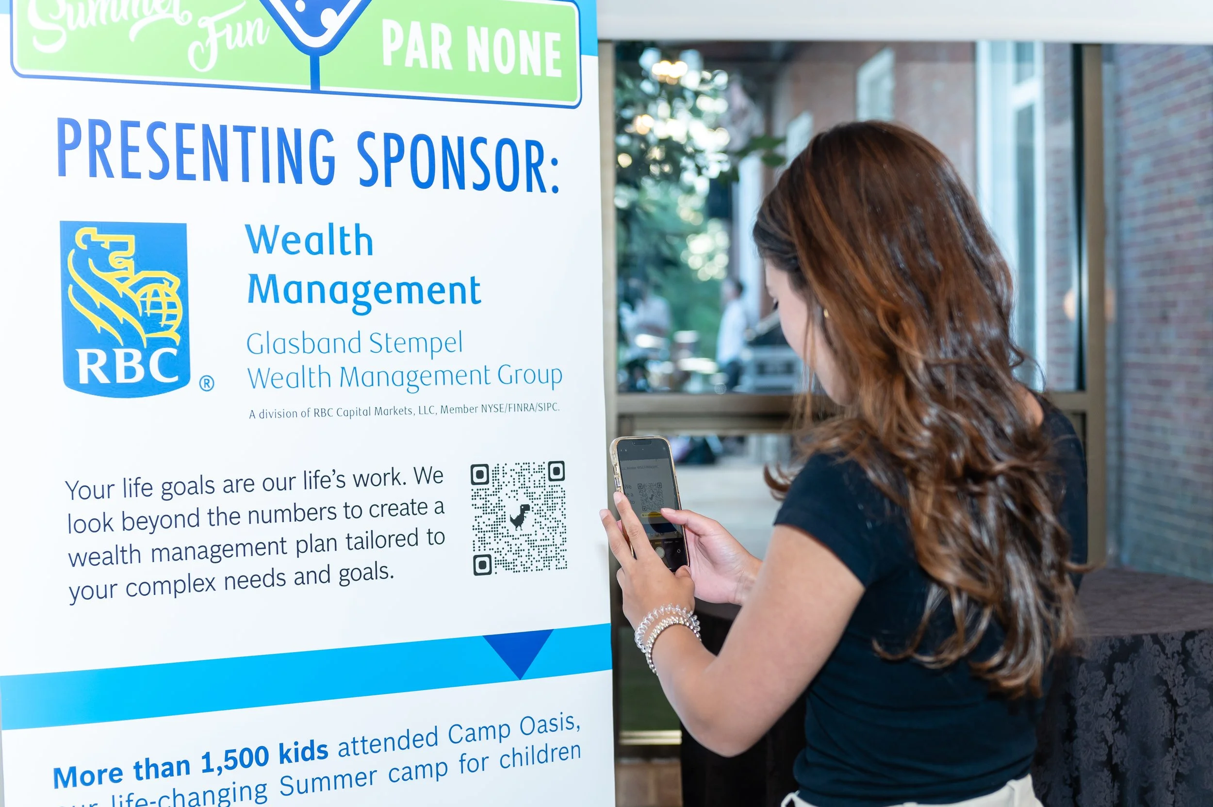 A woman with long, curly brown hair is interacting with a QR code on a large banner with her smartphone. The banner has the RBC sponsor logo on it and text about wealth management. The woman is wearing a black top and a pearl bracelet, standing indoo