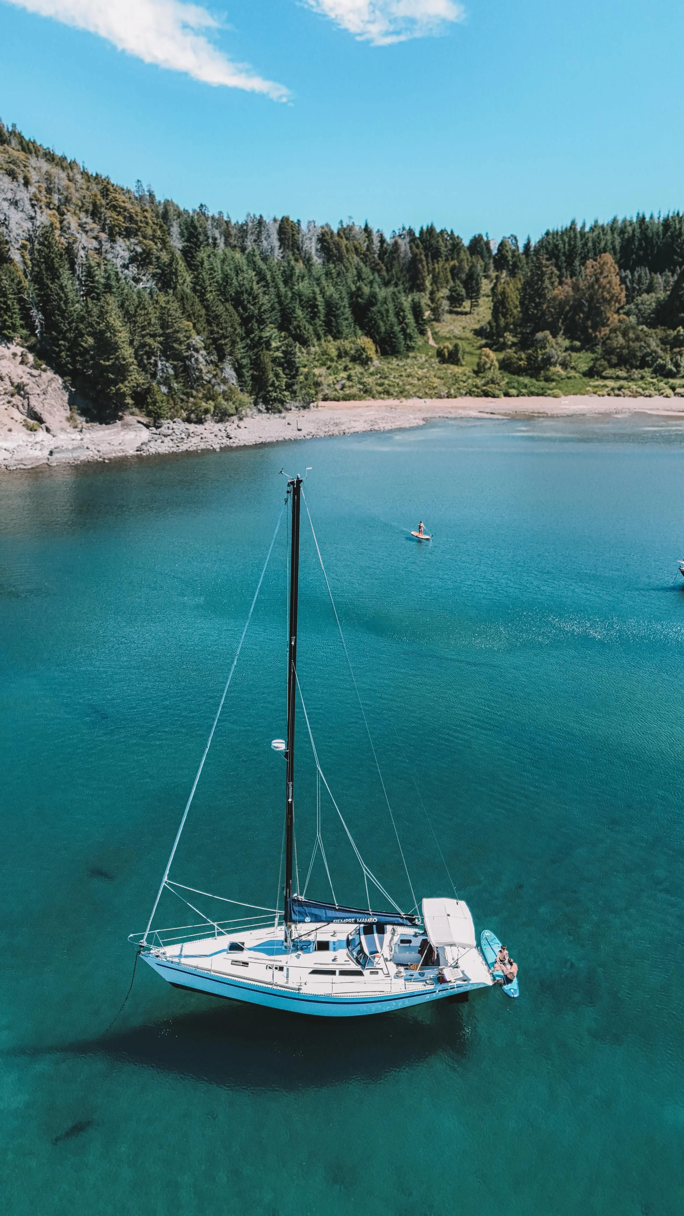 A sailboat floating on calm turquoise water near a forested shoreline with a person paddleboarding in the background.