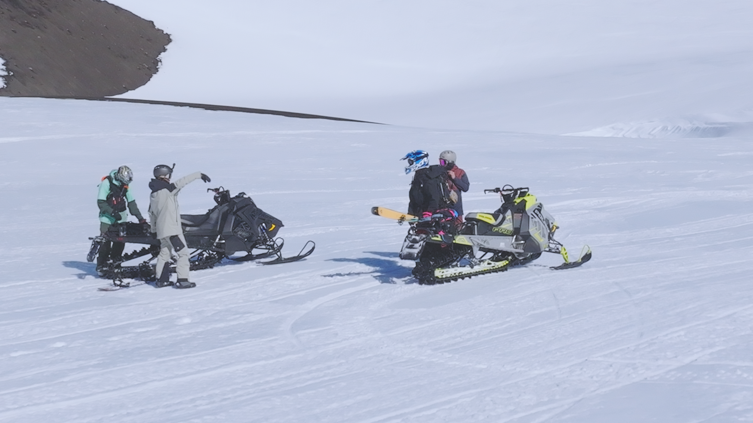 Four people in winter gear with helmets and goggles standing on a snowy landscape, with two snowmobiles parked nearby.
