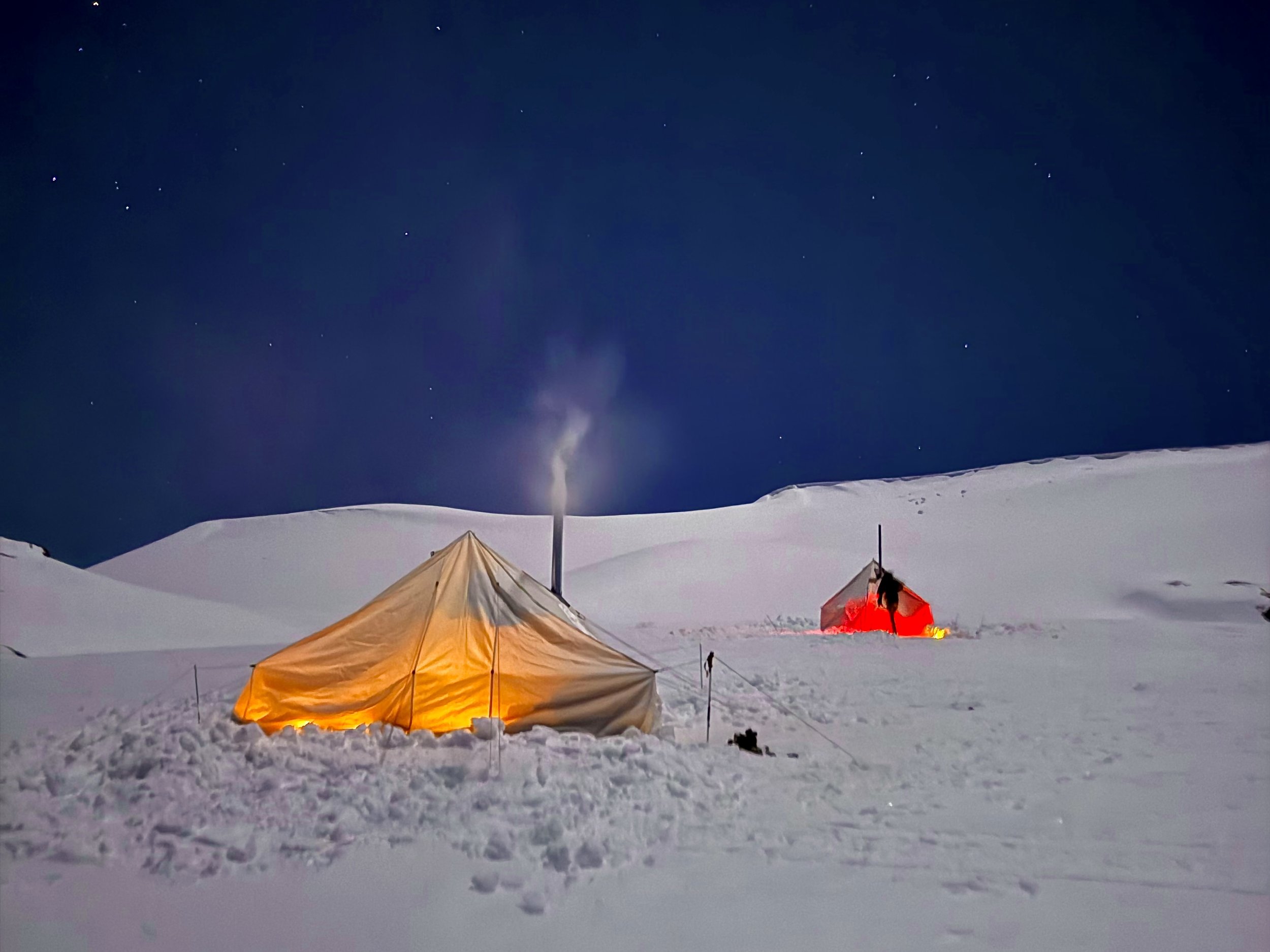 Snow-covered landscape at night with two tents, one glowing with yellow light and the other red, surrounded by snow, with a smokestack emitting smoke and stars visible in the dark sky.