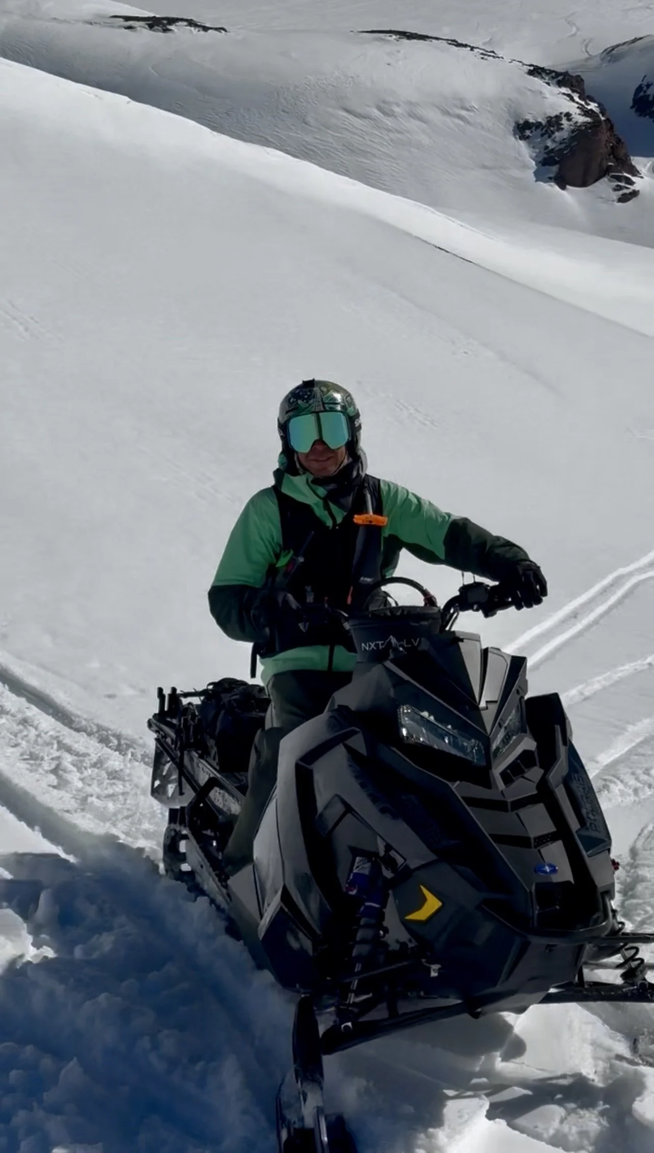 A person dressed in green and black winter gear, wearing a helmet and goggles, riding a snowmobile in a snowy mountainous landscape.
