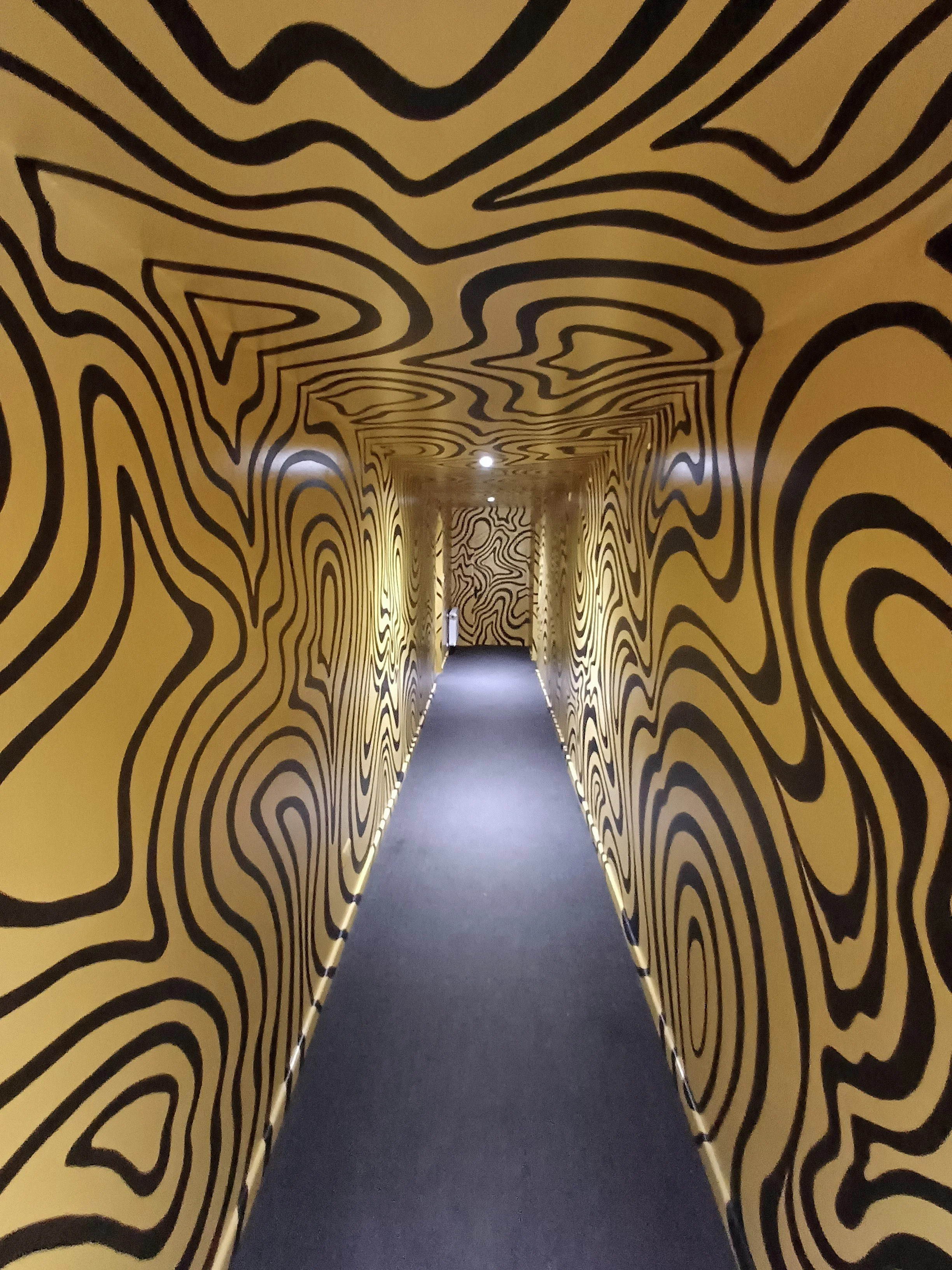 A narrow corridor with black line patterns on yellow walls and ceiling, creating an optical illusion effect.