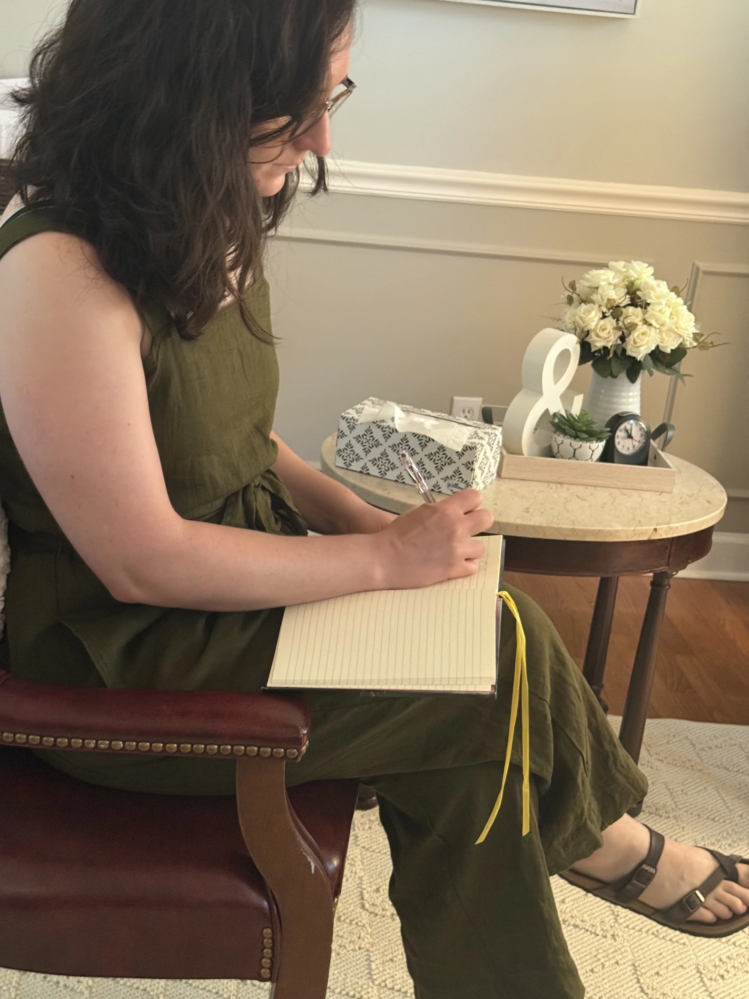 A woman with long curly hair and glasses sitting on a wooden chair with a red leather seat, writing in a yellow notepad. She is wearing a green dress and sandals. On a small round table next to her are a box of tissues, a white decorative sculpture, a bouquet of white roses, a small clock, and a potted plant.