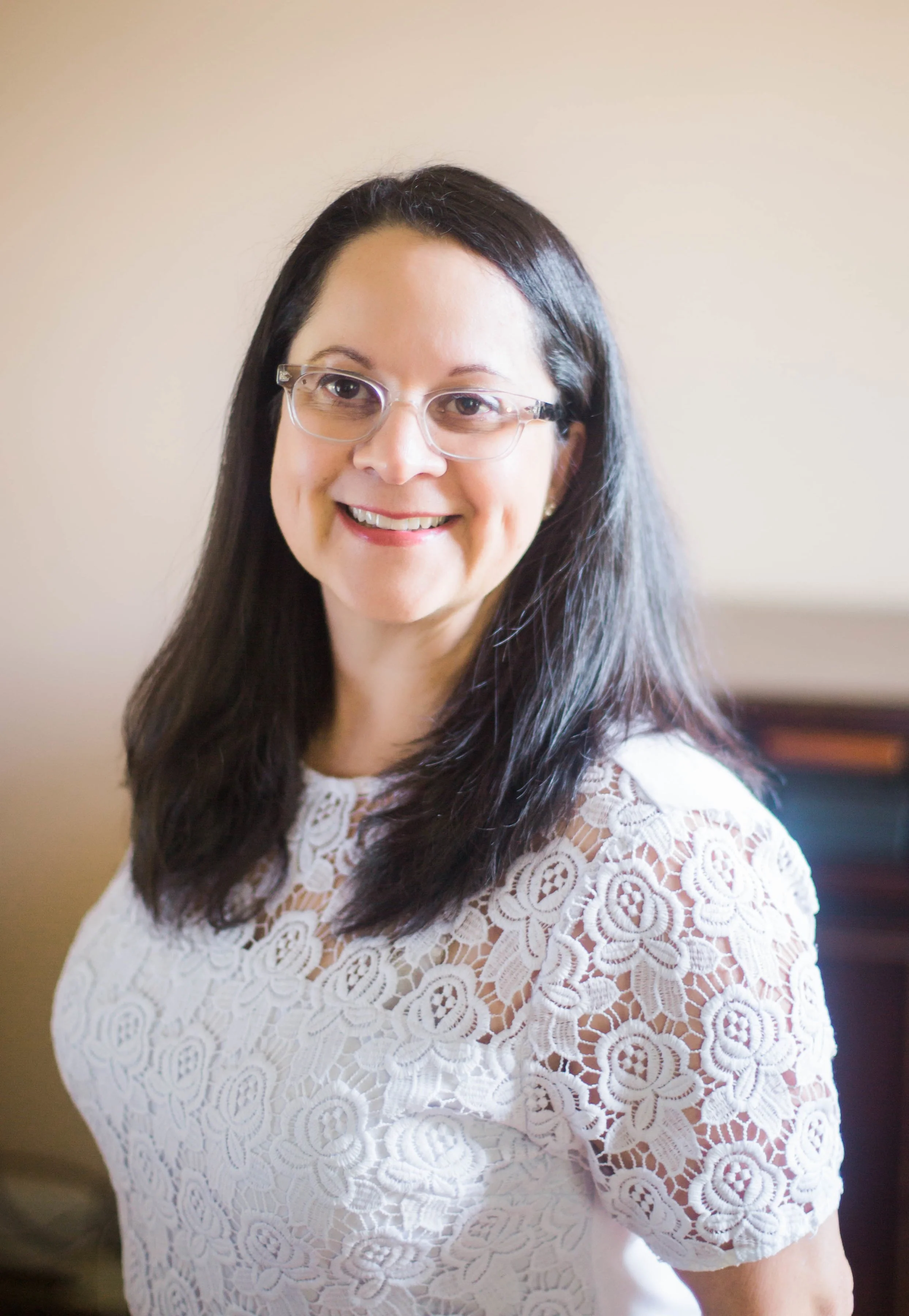 A woman with long dark hair and glasses, smiling, wearing a white lace top, standing indoors in front of a light-colored wall.