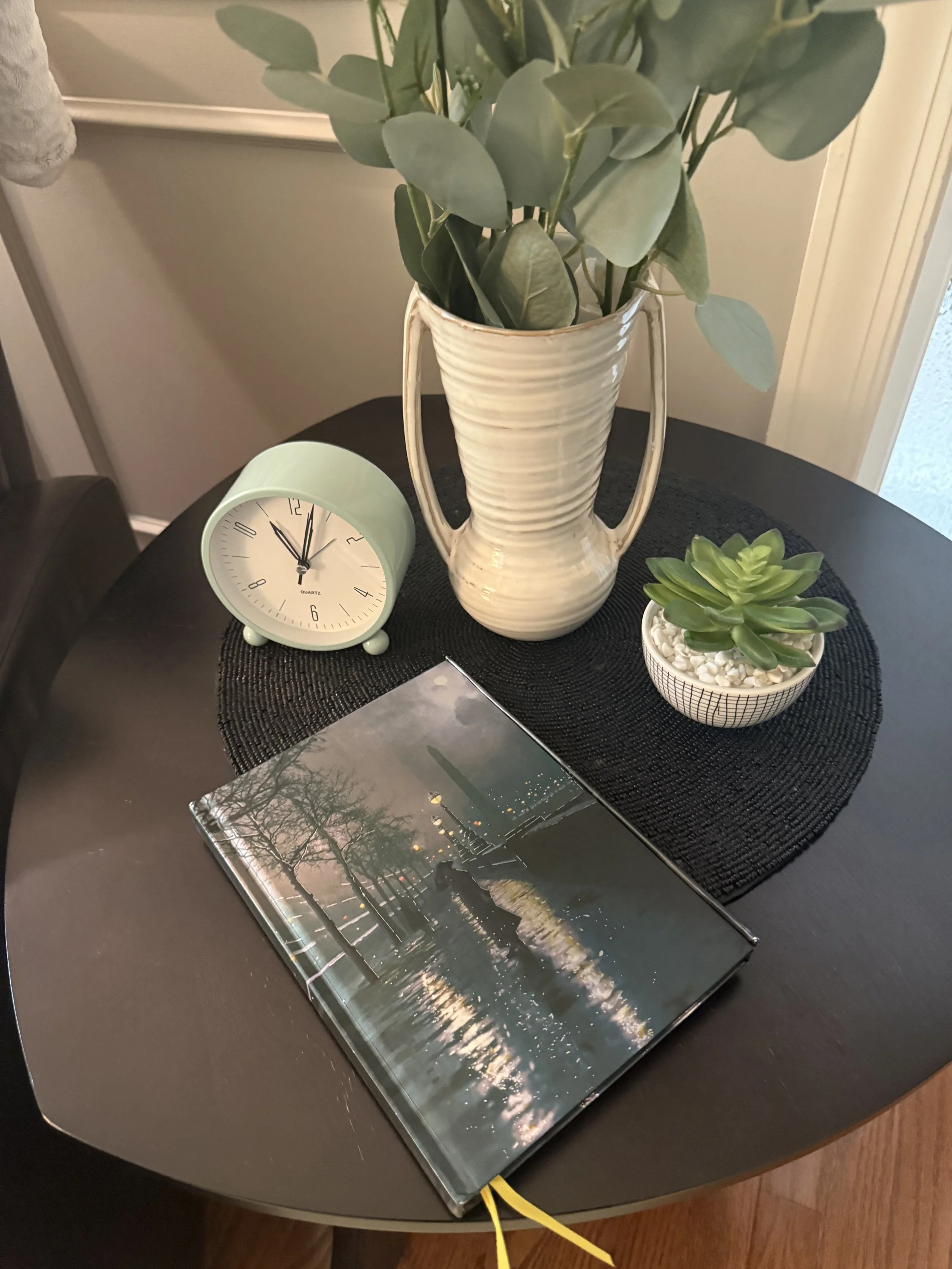 A round black table with a black woven placemat, a potted green succulent, a white vase with green leaves, a green alarm clock, and a photograph of a cityscape with a canal, trees, and a tower at sunset.