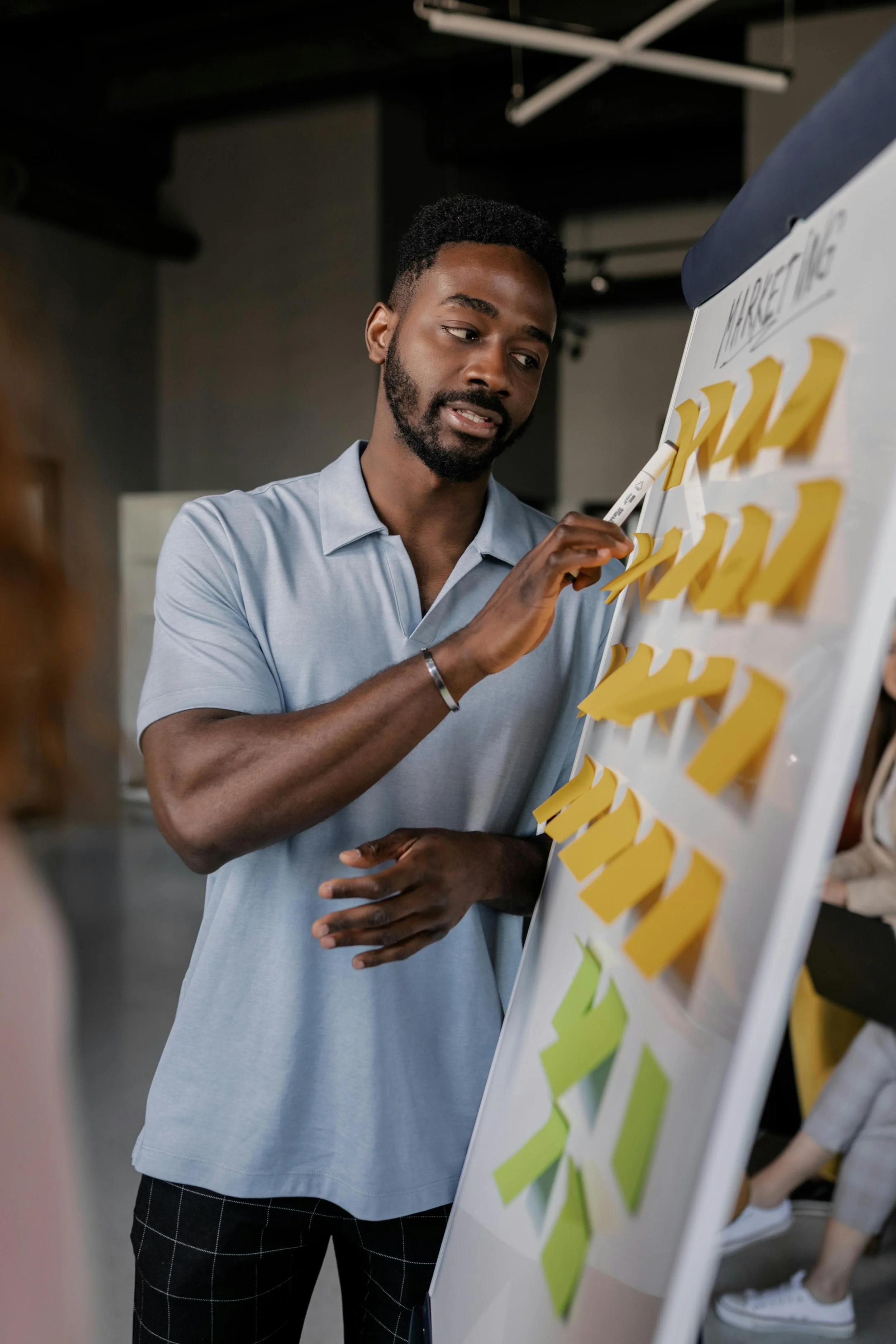 A young man in a light blue polo shirt stands next to a whiteboard covered with yellow and green sticky notes, actively participating in a planning session or workshop.
