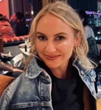 Close-up of a smiling young ADHD woman with curly hair wearing glasses, hoop earrings, and layered necklaces, indoors with a blurred background.