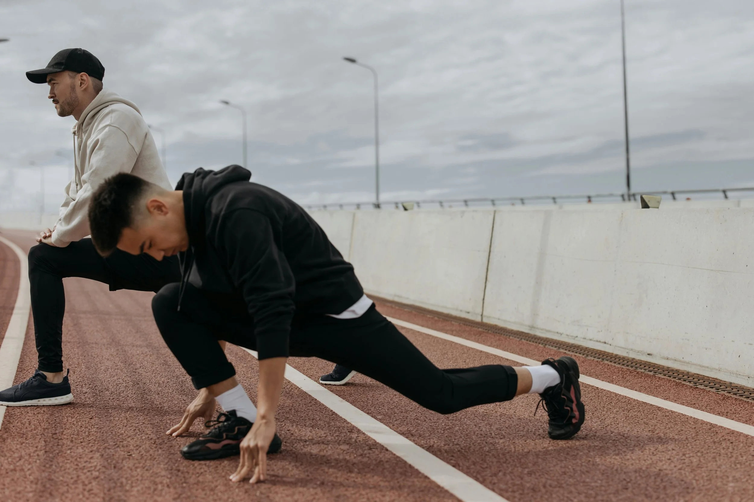 Two men in athletic clothing doing warm-up exercises on a red outdoor running track, with a cloudy sky overhead.