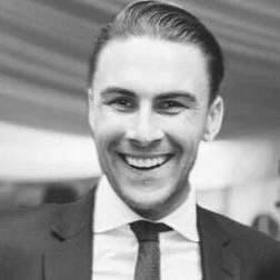 Young man smiling, wearing a suit and tie, with a blurred background.