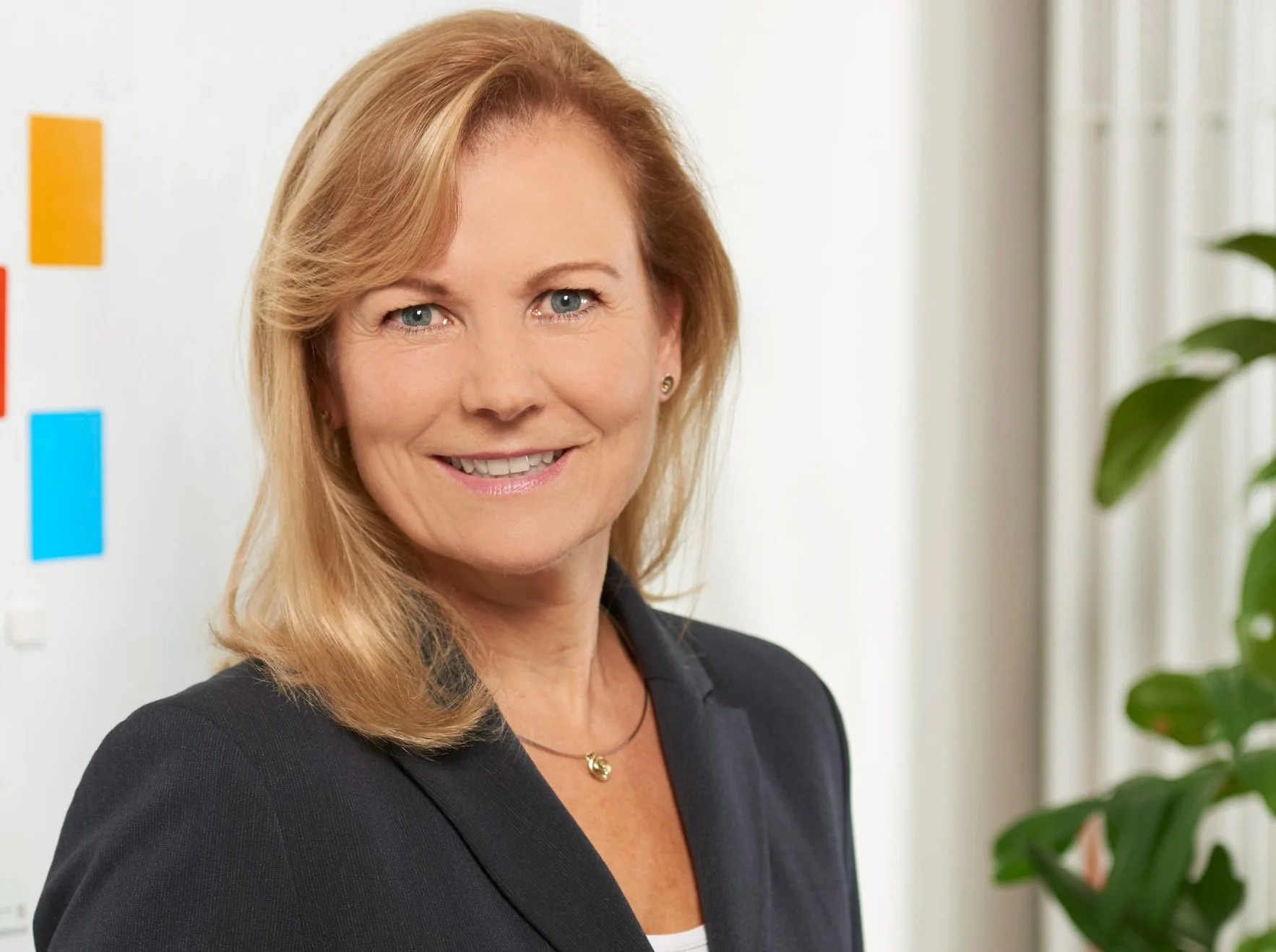 Porträt einer lachenden Frau in einem schwarzen Blazer in einem Büro, mit bunten Post-its an der Weißwand im Hintergrund und einem grünen Pflanzen an der Seite.