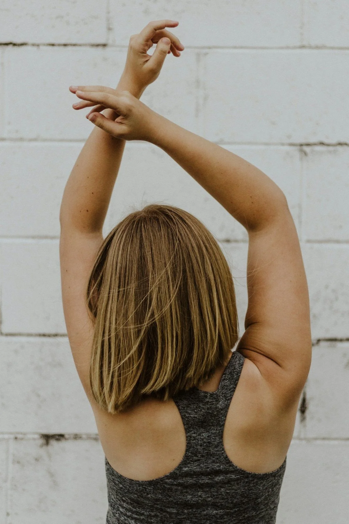 Nahaufnahme einer jungen Frau mit braunen, glatten Haaren, die den Rücken zeigt. Sie trägt ein graues Sporttop und hebt die Arme über den Kopf, während sie sich streckt oder tanzt, vor einer weißen Ziegelwand.